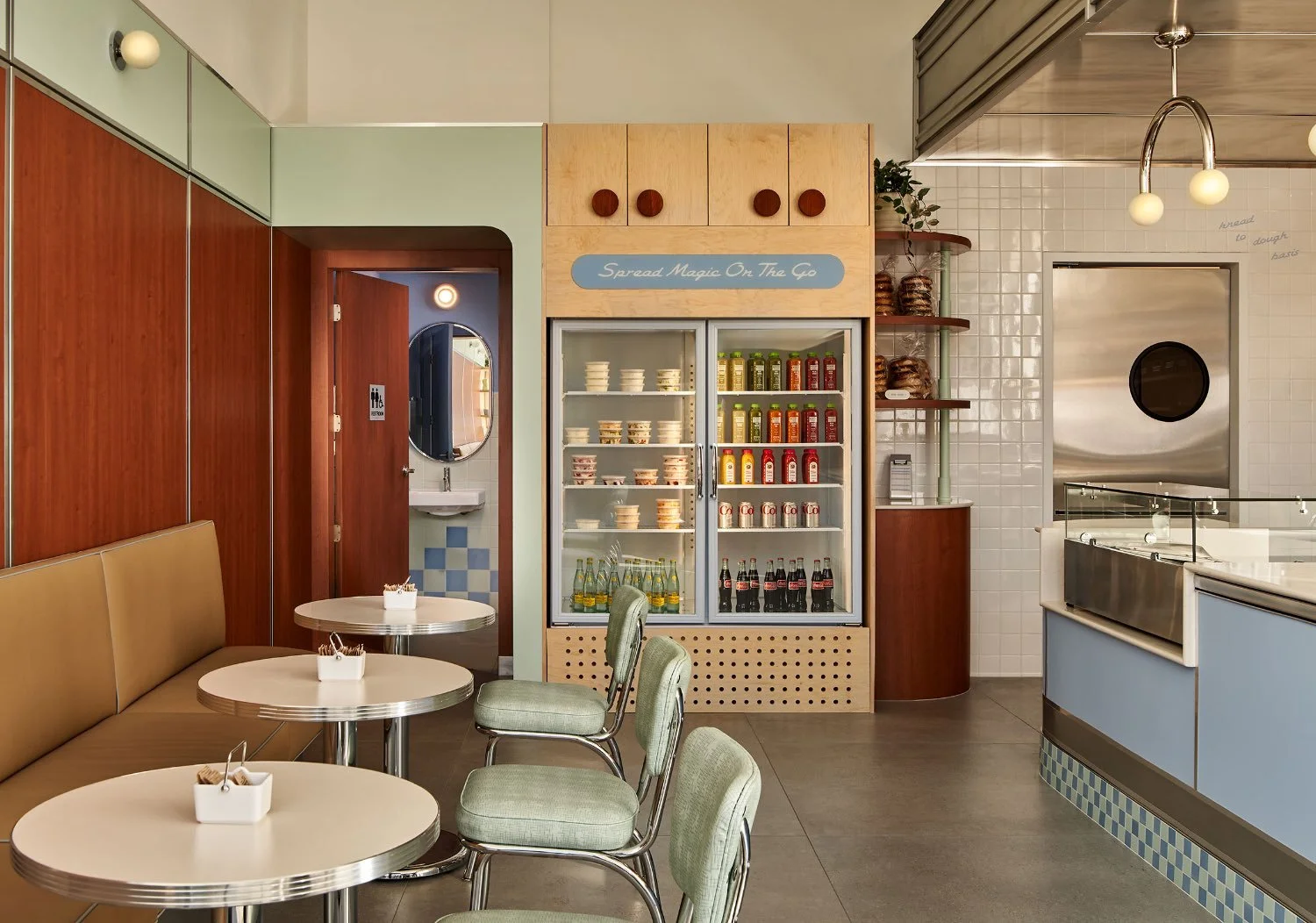 Inside a vintage-style cafe with round tables, green chairs, a refrigerator with assorted drinks, a seating area with a beige bench, a door to a bathroom, and a counter with baked goods and a small glass display.
