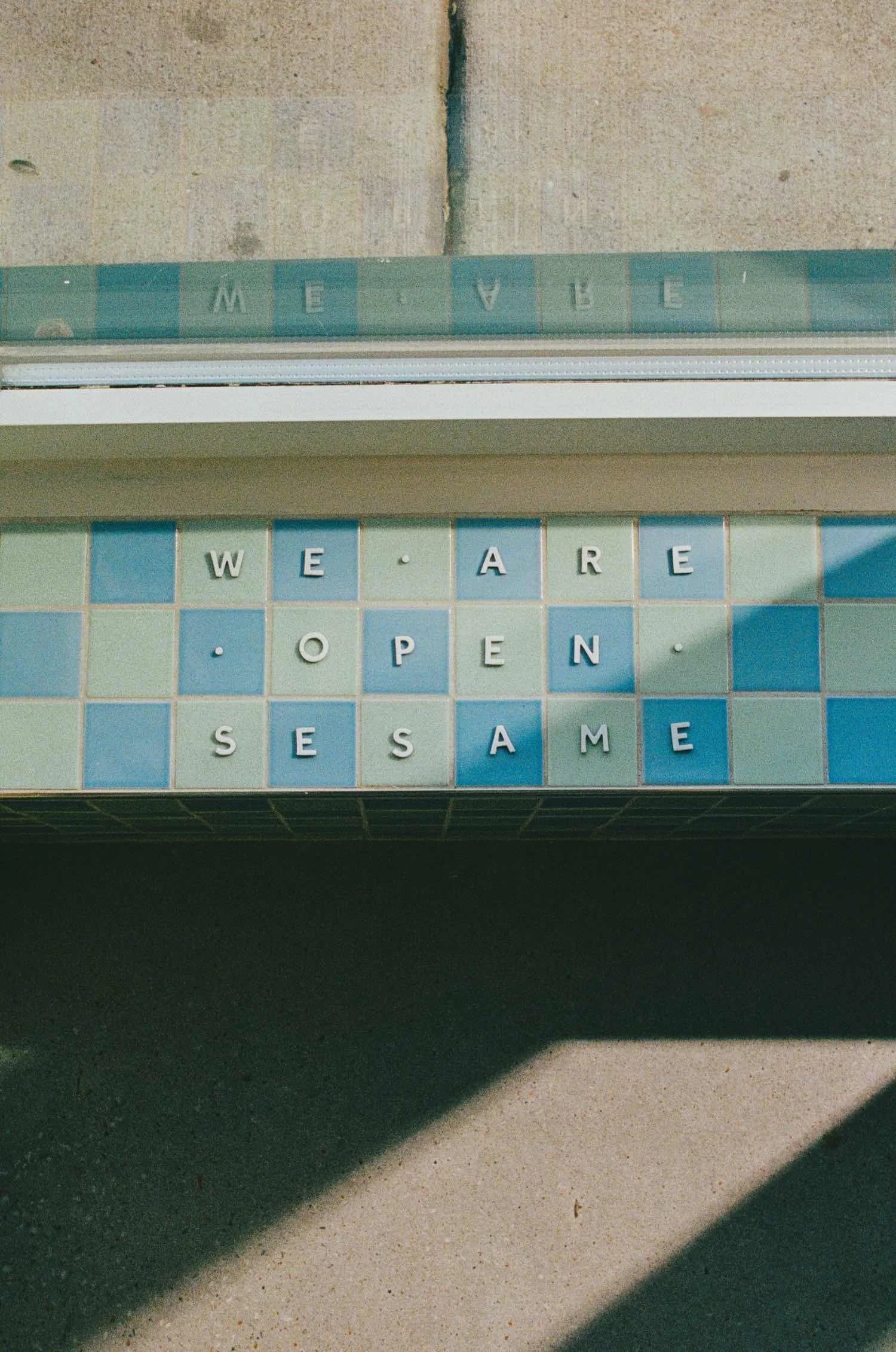 A daytime photo of a storefront with a sign made of tiles that spell out 'WE ARE OPEN.' The sign is mounted above the entrance, with shadows cast on the sidewalk below.