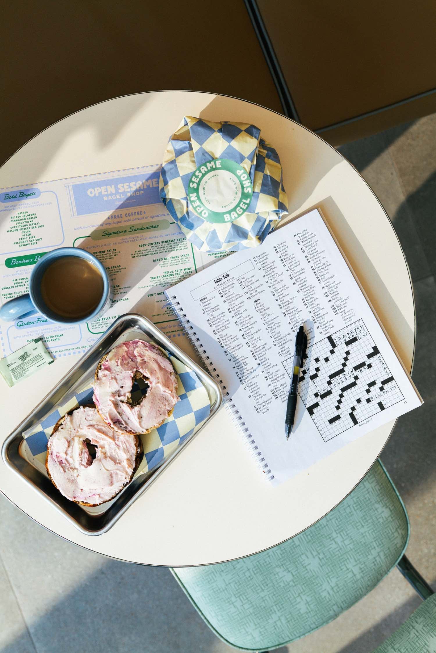 A table with a sliced ice cream sundae, a cup of coffee, a wrapped bagel in branded paper, a menu, a crossword puzzle, and a pen.