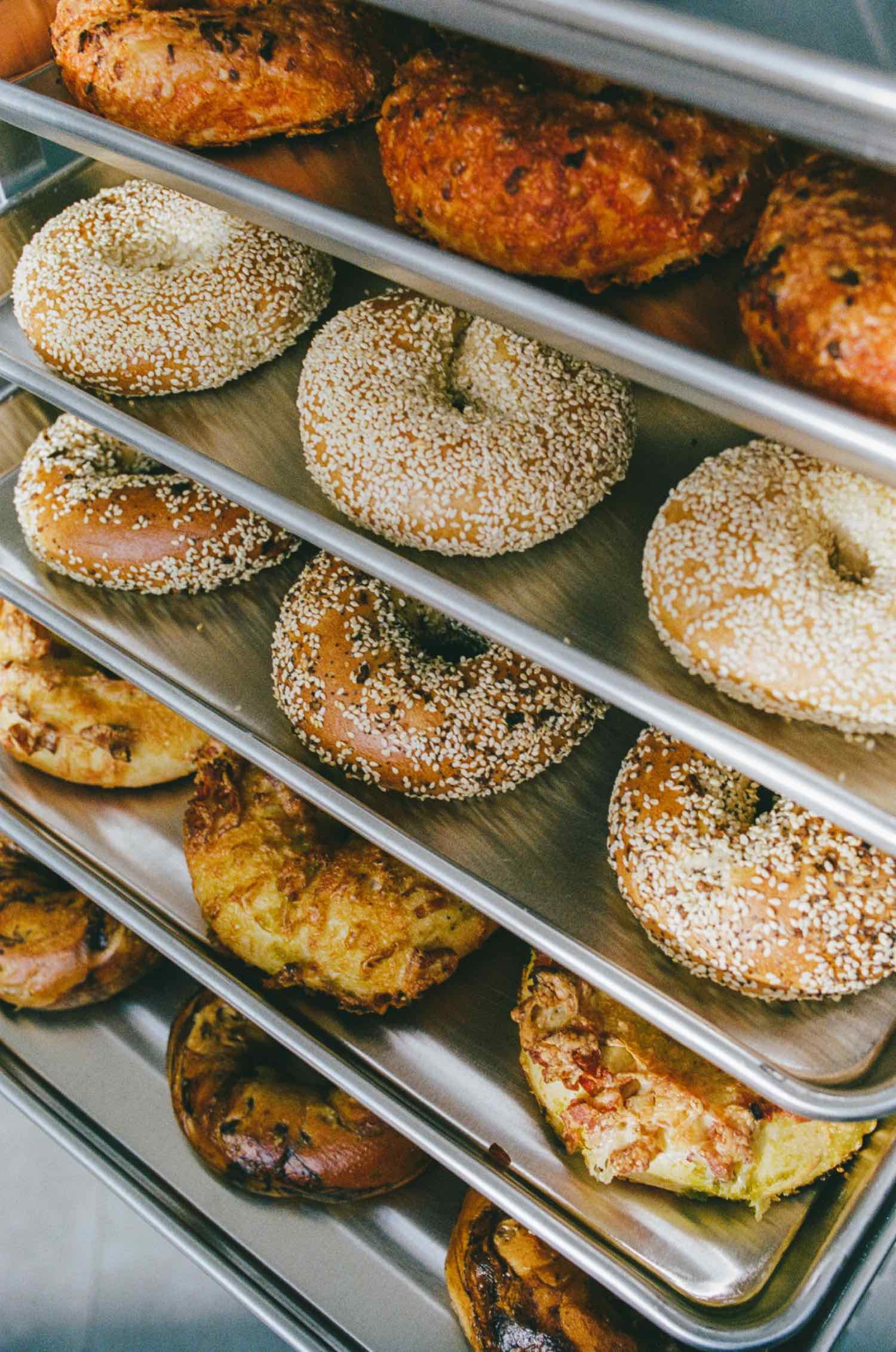 Multiple trays of baked bread and pizza slices, including sesame seed bagels and pizza with toppings.