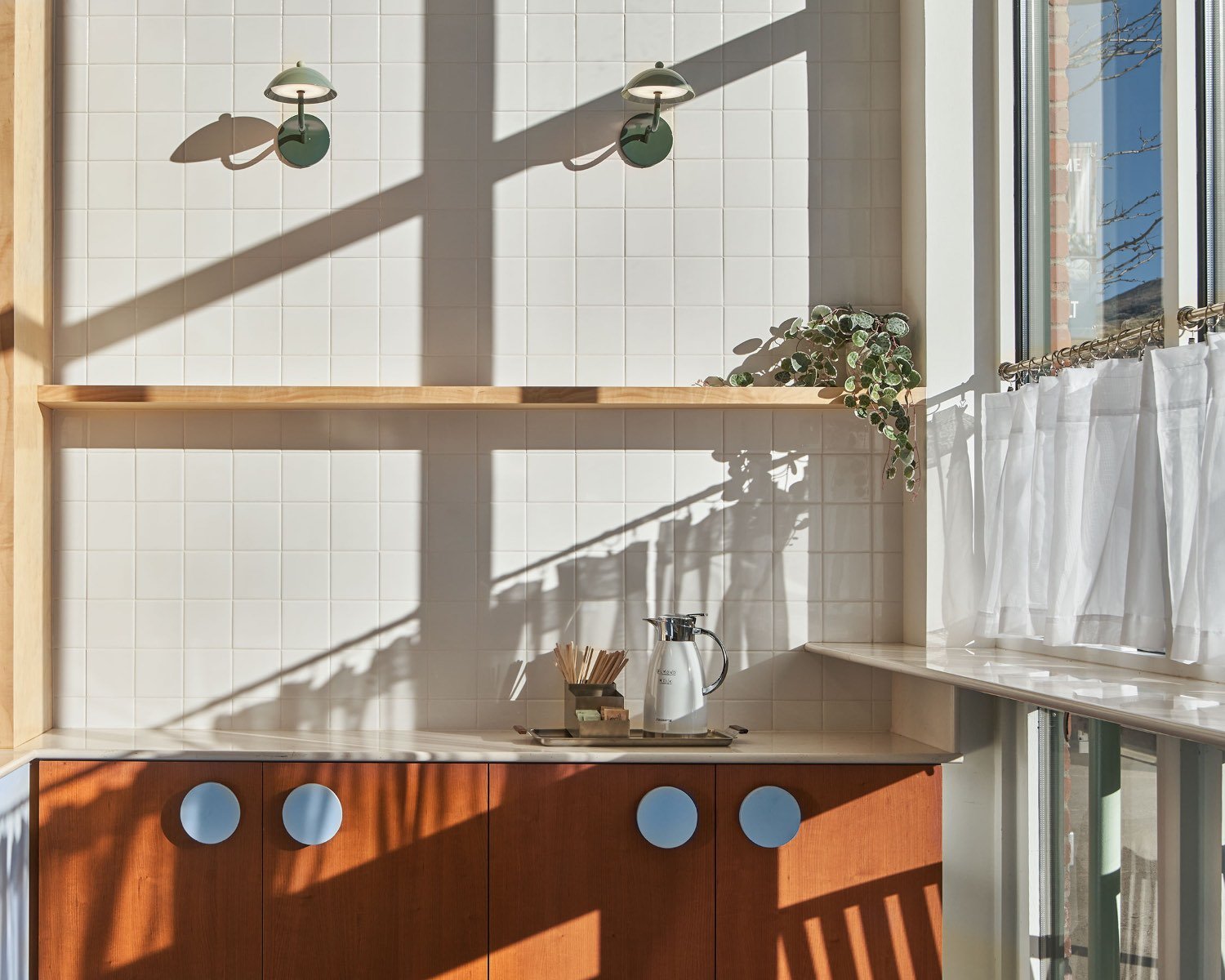 A section of a kitchen with white tiled walls, sunlight casting shadows, a wooden cabinet with circular cutouts, a silver pitcher, a small container with wooden stirrers, a small plant, a window with white curtains, and two wall-mounted lamps.