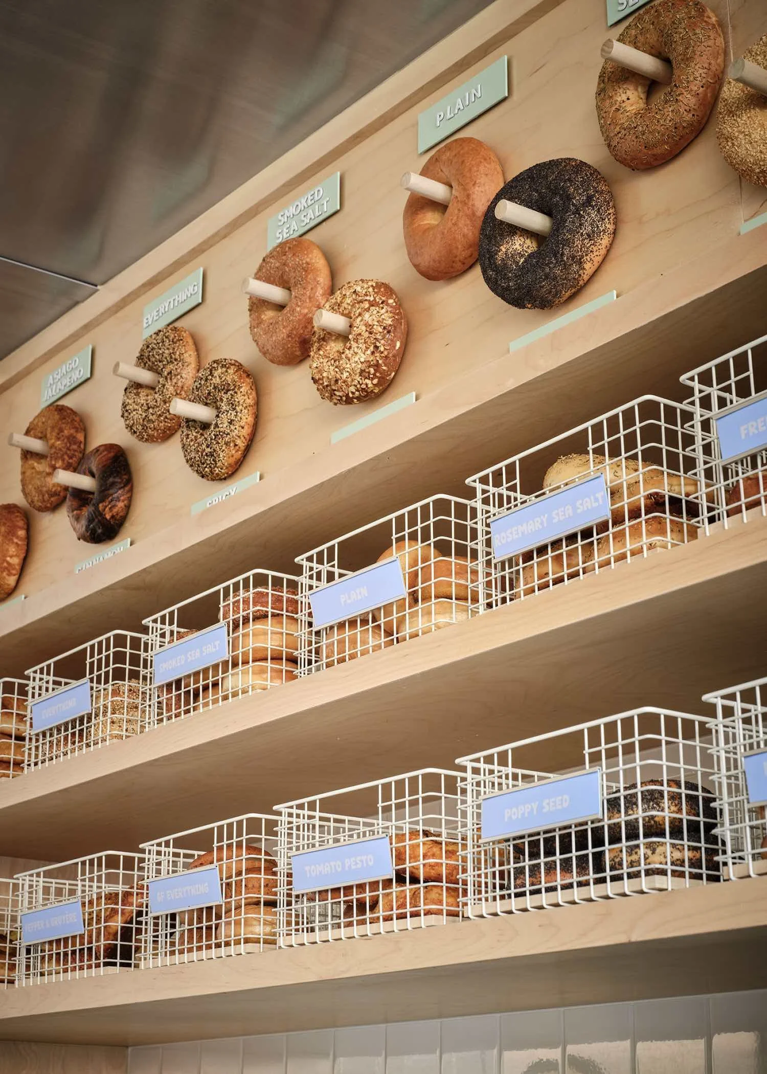 Display of various bagels arranged on wooden shelves labeled with different flavors and toppings such as plain, smoked sea salt, everything, poppy seed, tomato pesto, and sesame seed in a bakery or bagel shop.