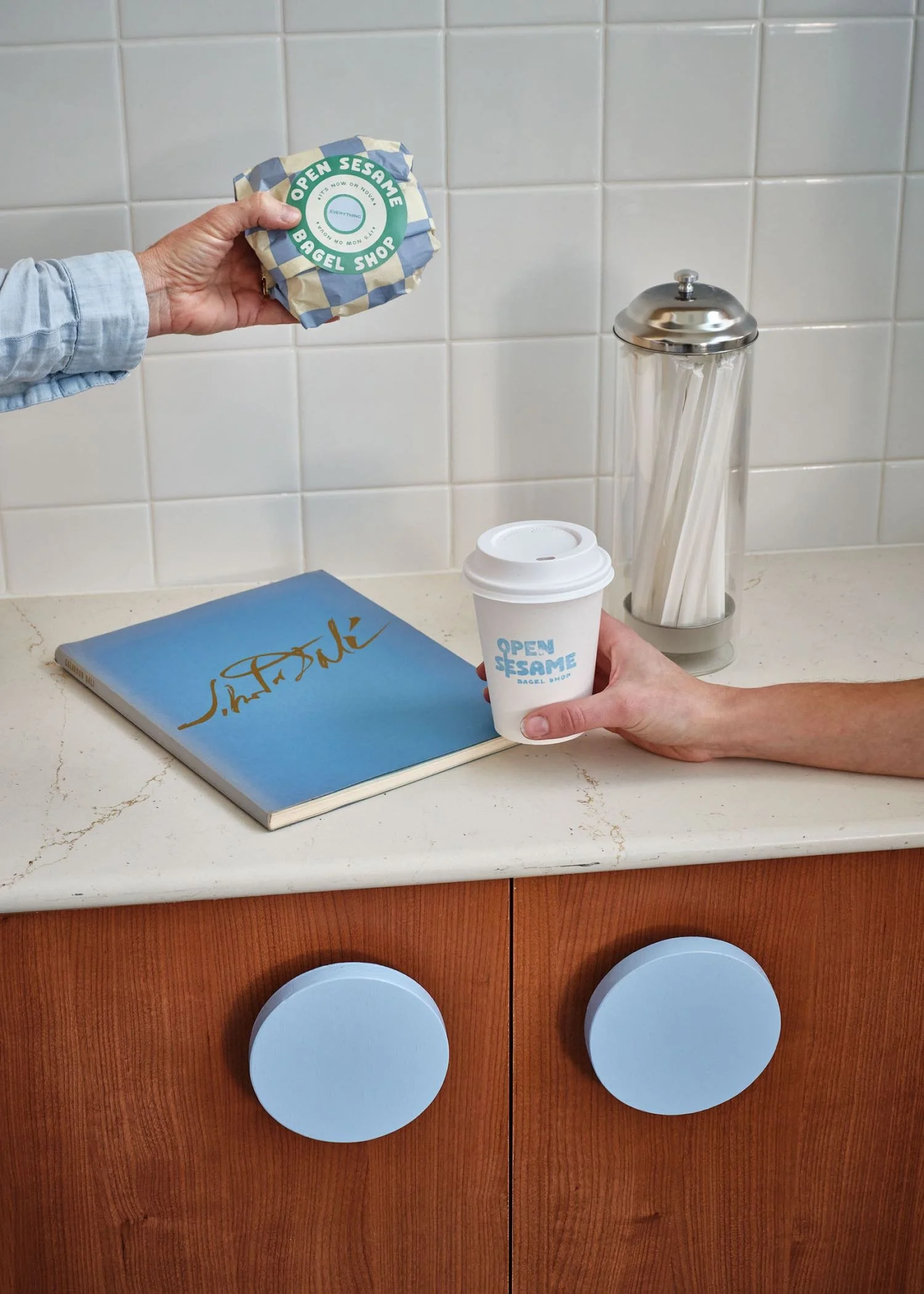 Hand holding a wrapped bagel from Open Sesame Bagel Shop, with a coffee cup, a book, and a napkin on a kitchen counter.