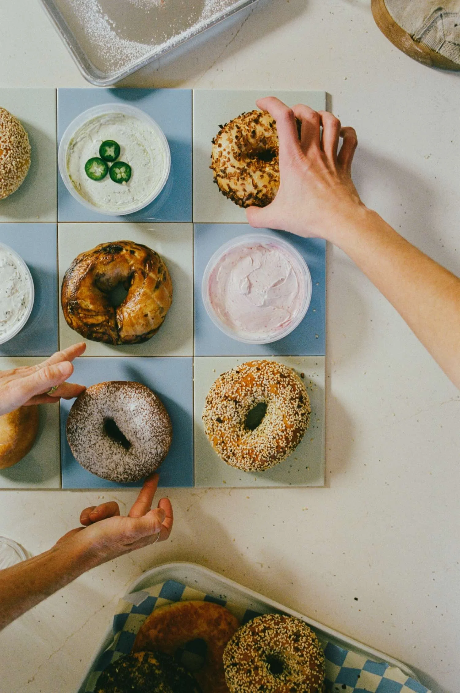 A top-down view of a table with various bagels and cream cheese spreads, some with toppings like sliced jalapenos and sesame seeds. Hands are reaching for or pointing at the bagels.