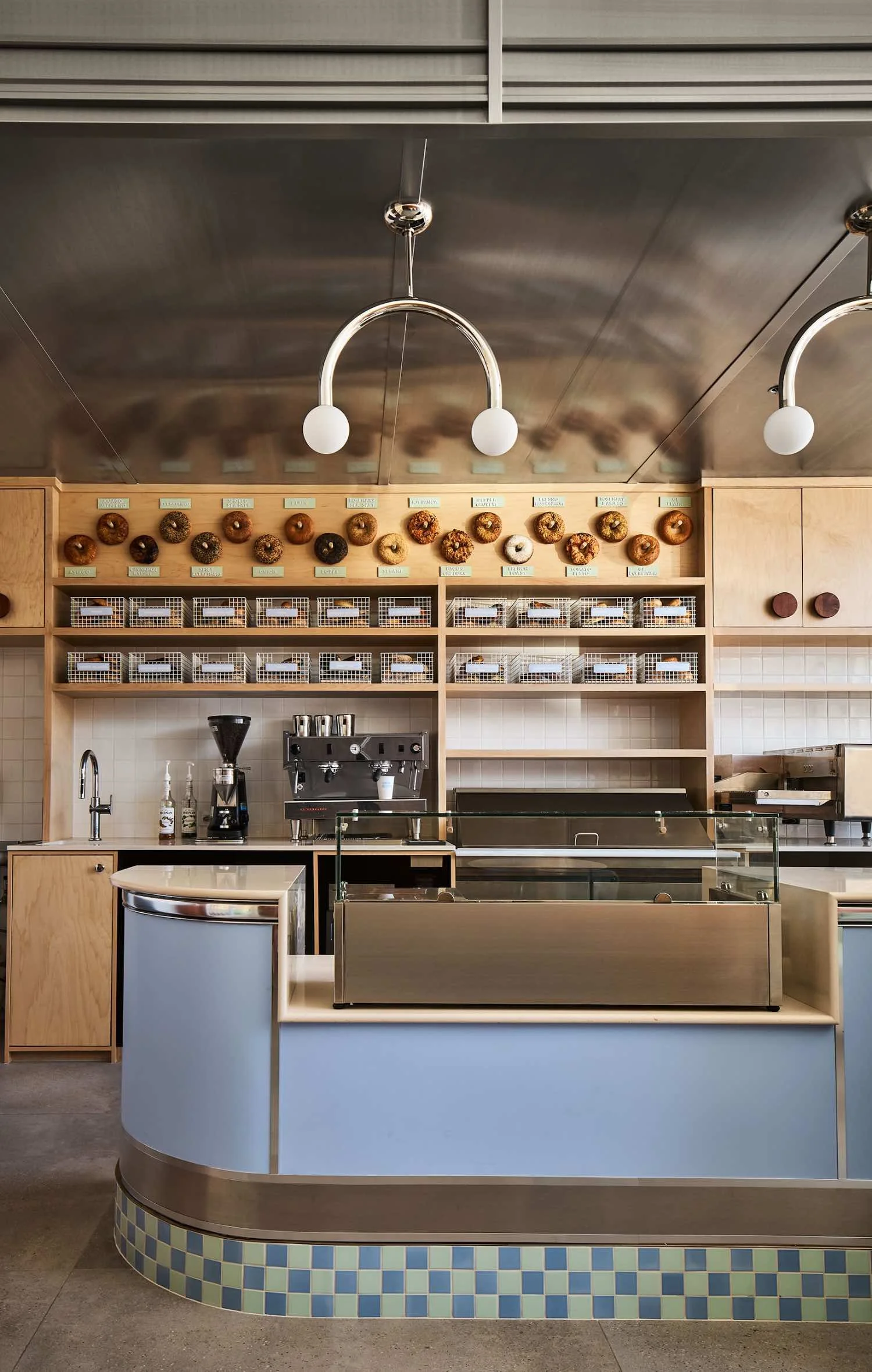 Interior of a modern coffee shop or bakery with a light blue and beige counter, wooden shelving with donut displays, and a metal ceiling with modern white light fixtures.