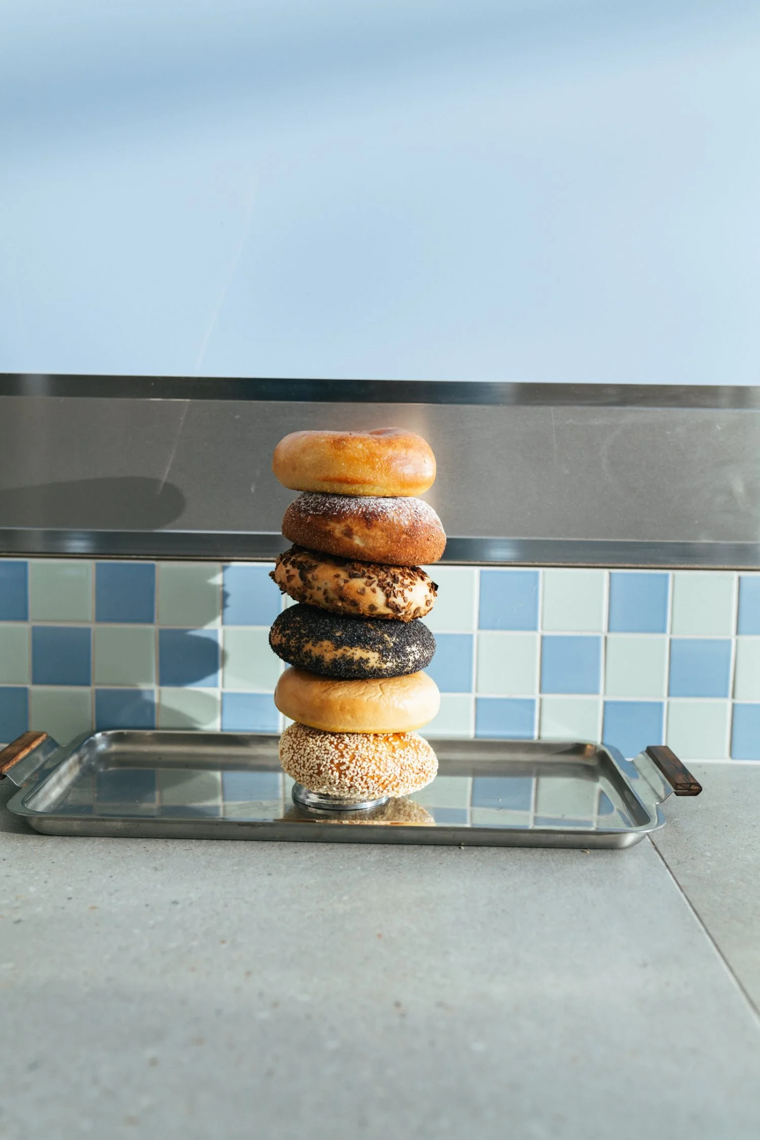 Stack of nine assorted donuts on a metal tray in front of a blue tiled wall.