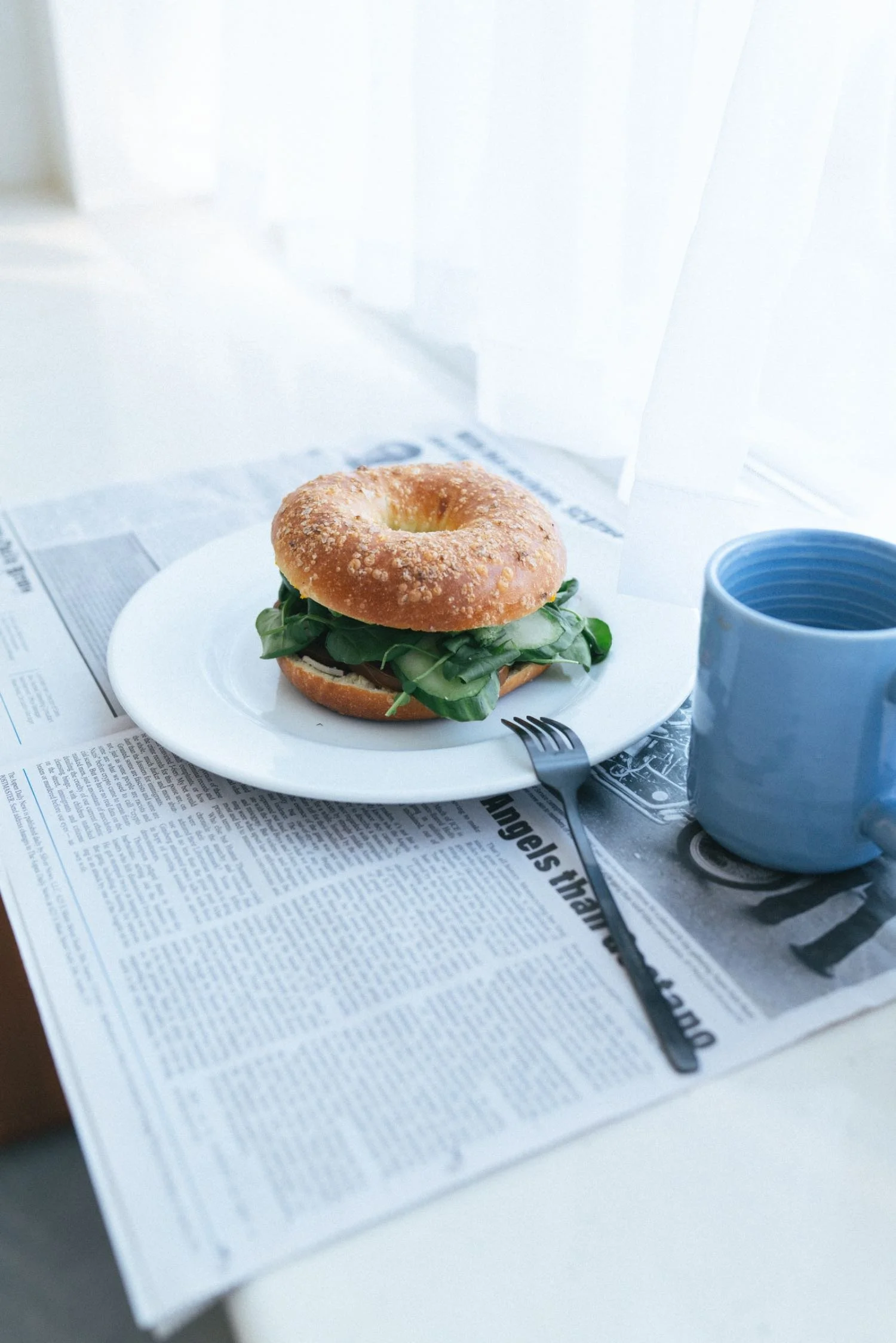 A plate with a bagel sandwich containing spinach and other ingredients, a fork, a mug, and a newspaper on a table.