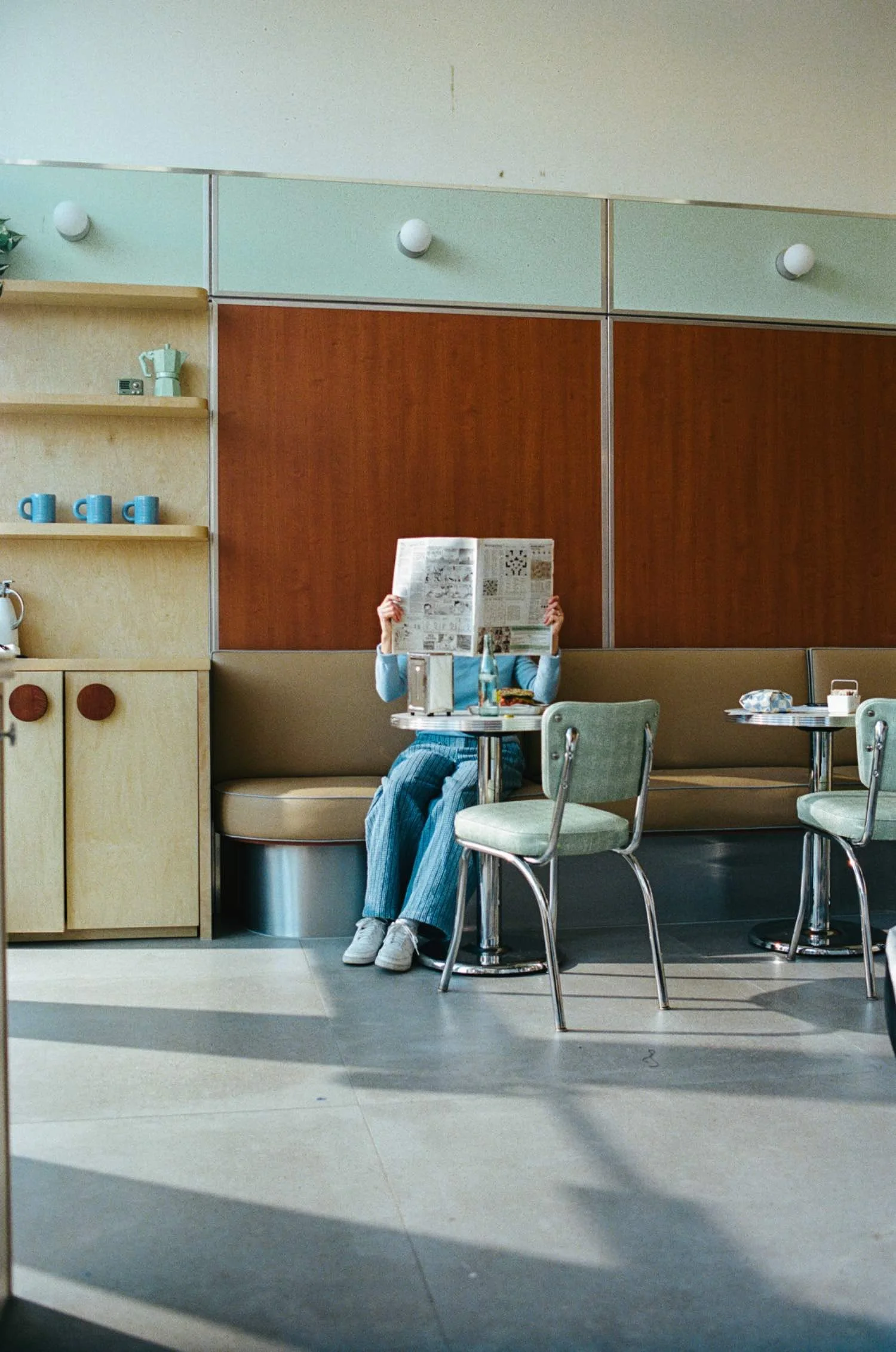 Person sitting at a table in a retro-style diner or cafe, reading a newspaper with legs crossed. The setting includes vintage chairs, a wooden bench, and a wood-paneled wall with shelves holding mugs. The person is wearing light-colored sneakers, striped pants, and a long sleeve top.