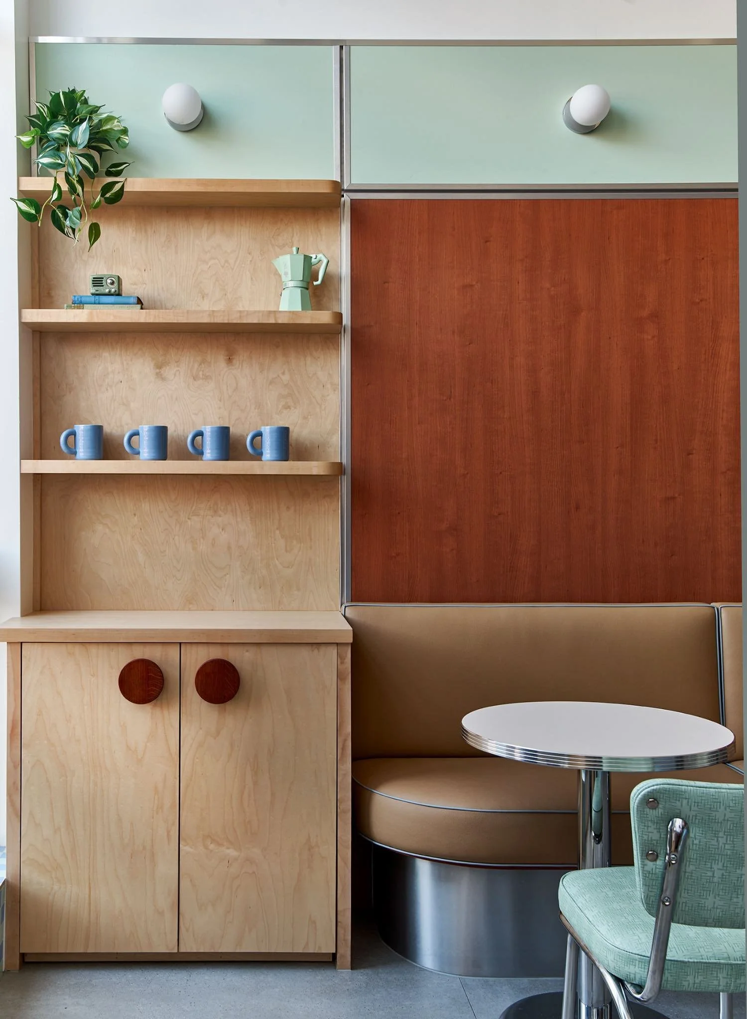 Interior of a retro-style diner with a wooden cabinet, a small round table with a green chair, and a wall with pastel green and red panels. There are decorative mugs and a coffee pot on the shelves.