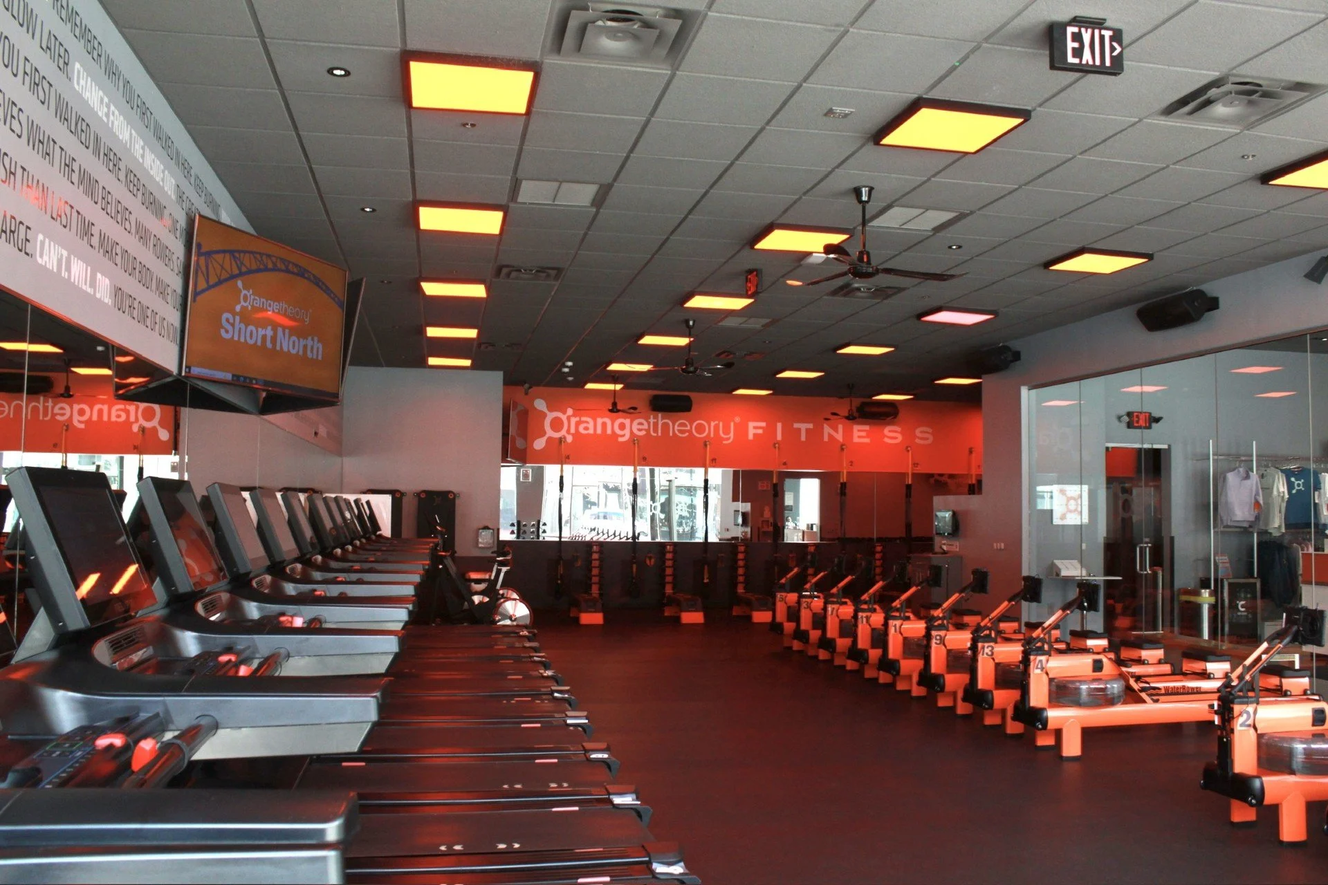 A CM Collaboration design of an Orange Theory gym with treadmills on the left and rowing machines on the right, lit by orange overhead lights.