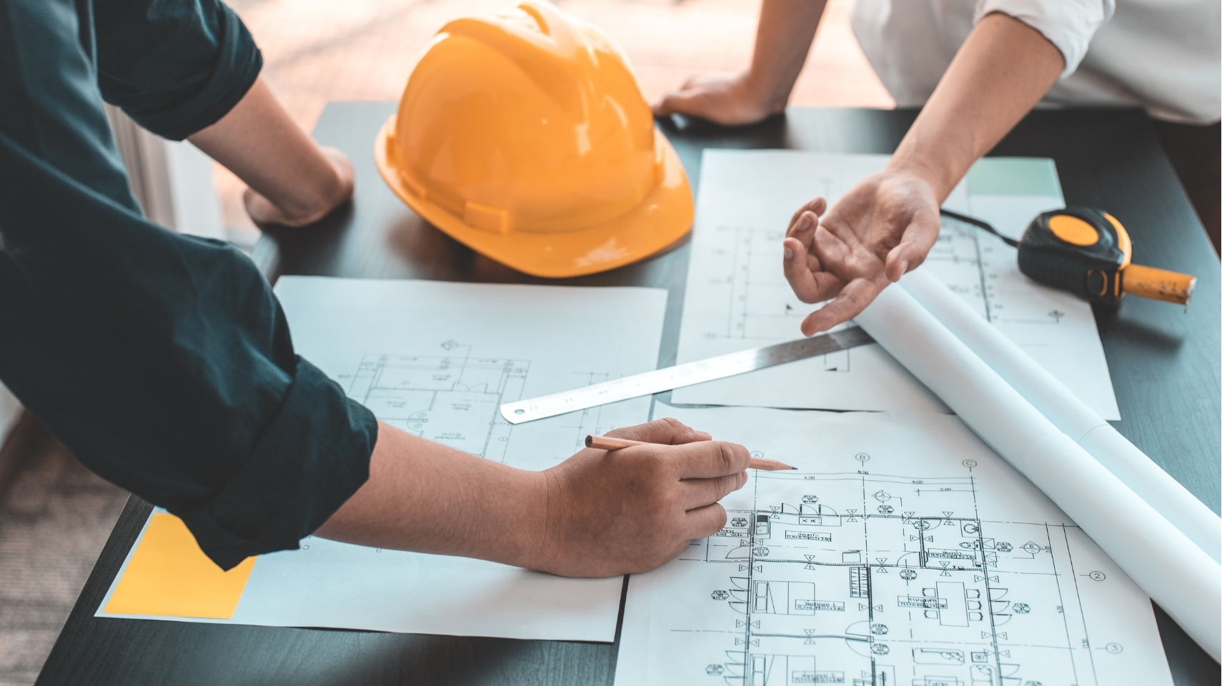 Two people in a commercial construction company review architectural blueprints on a table with a hard hat, ruler, and tape measure nearby.