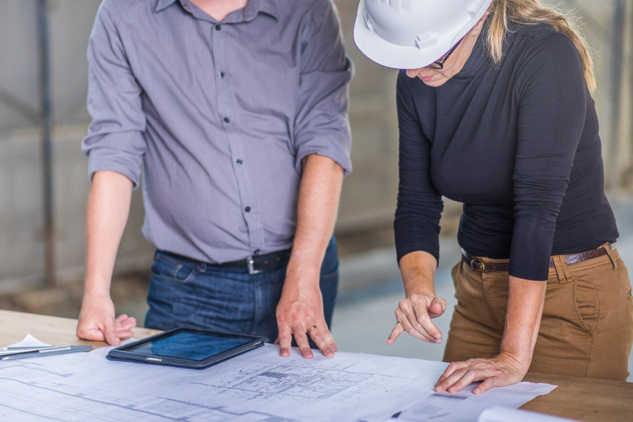 Two people review architectural blueprints on a table for a Construction consulting firm, CM Collaborative; one wears a hard hat and points at the plans.