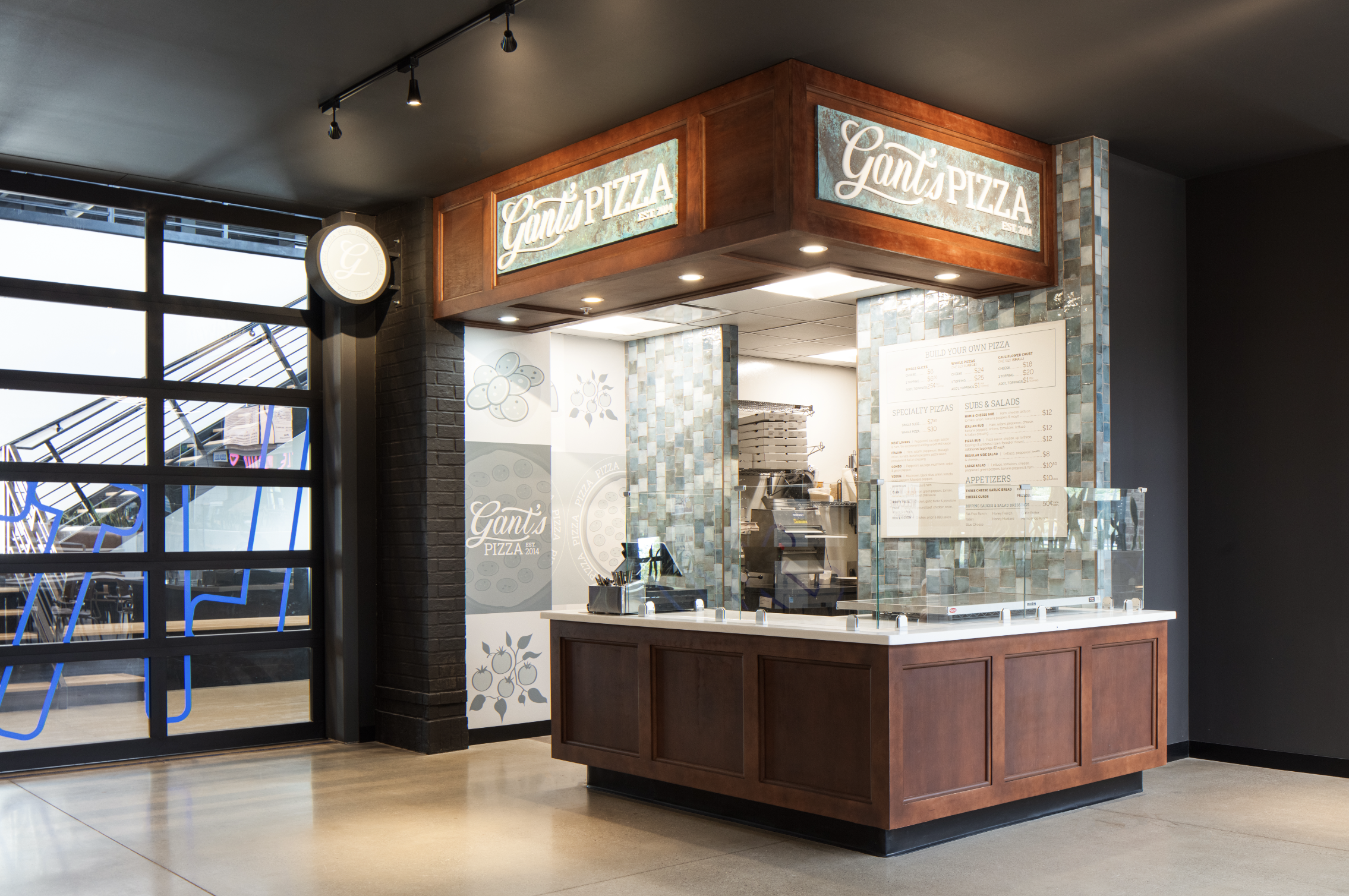 Small pizza counter named "Gani’s Pizza" with a menu board, glass tiles, and wood paneling in a modern building proving CM Collaboration's build-out project management.