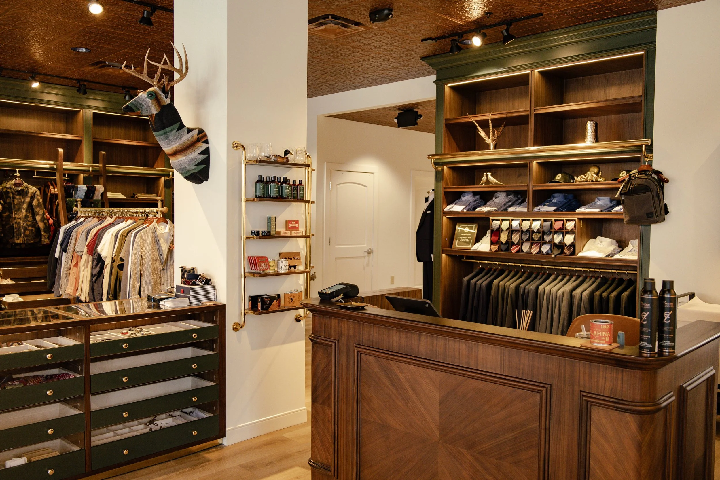 Men’s clothing store construction done by CM Collaborative, with suits, shirts, and accessories displayed near a wooden counter and open shelving.
