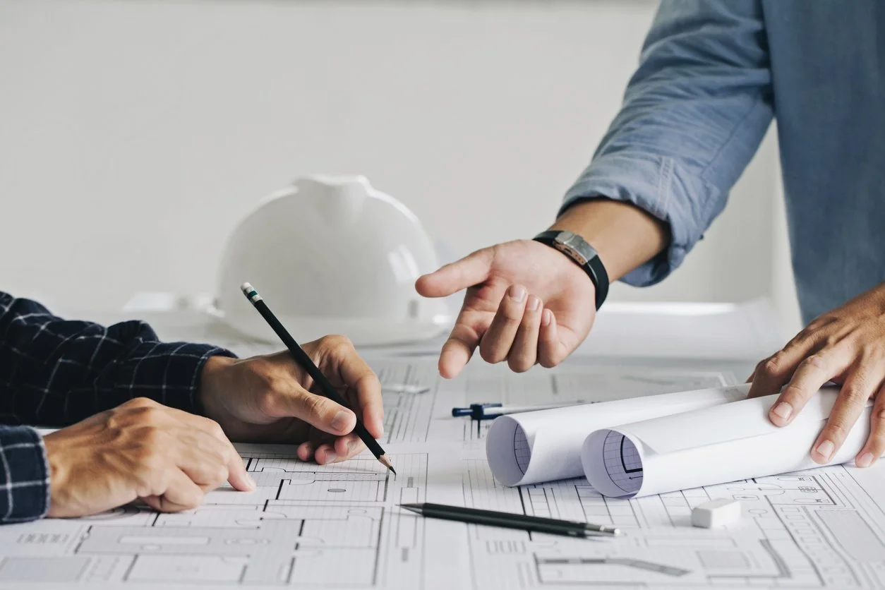 Two people discuss architectural blueprints on a desk with rolled plans, pens, and a white safety helmet visible showing the full lifecycle construction project management