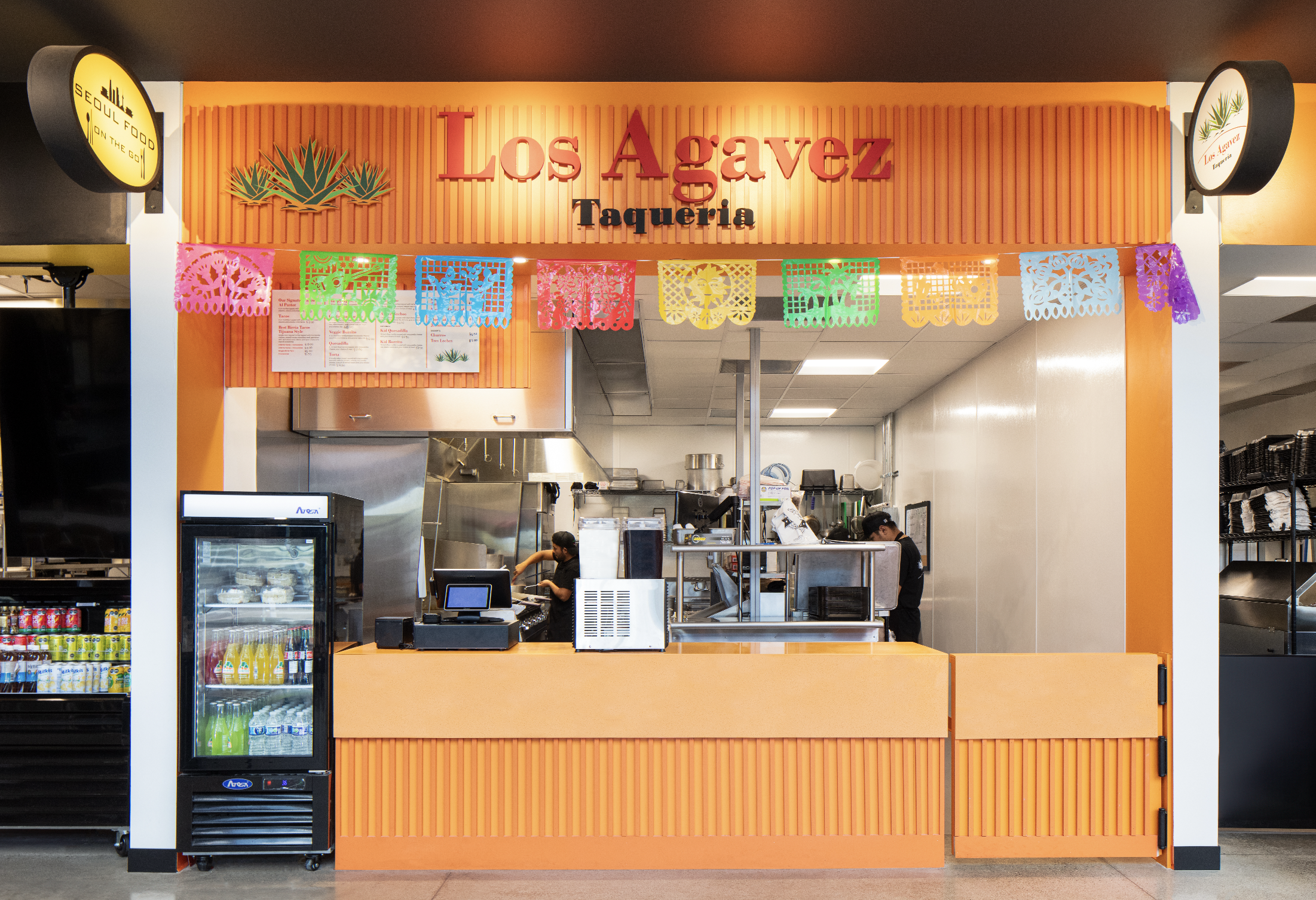 Front view of Los Agavez Taqueria counter with colorful papel picado and a visible fridge stocked with drinks done by construction budgeting experts.