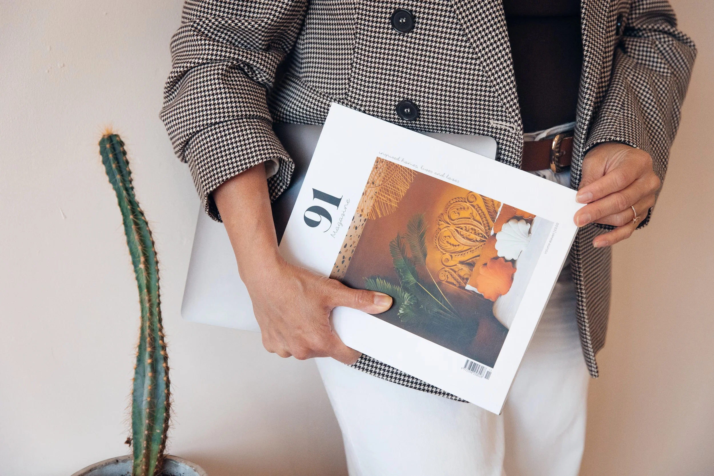 Person holding a magazine and a laptop, wearing a houndstooth blazer, with a tall cactus plant beside them.