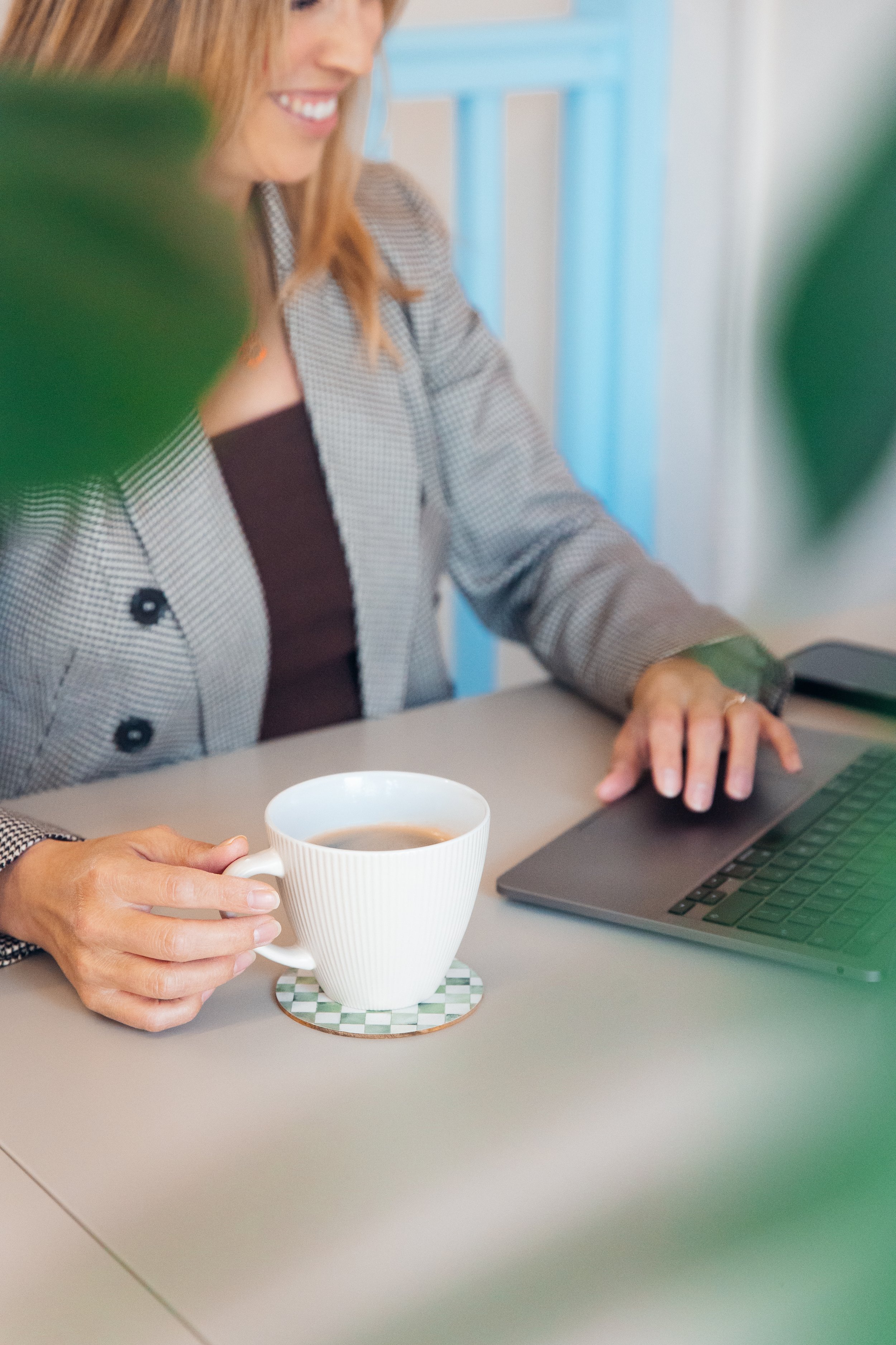 A woman smiling while working on a laptop at a desk, holding a mug of coffee or tea.