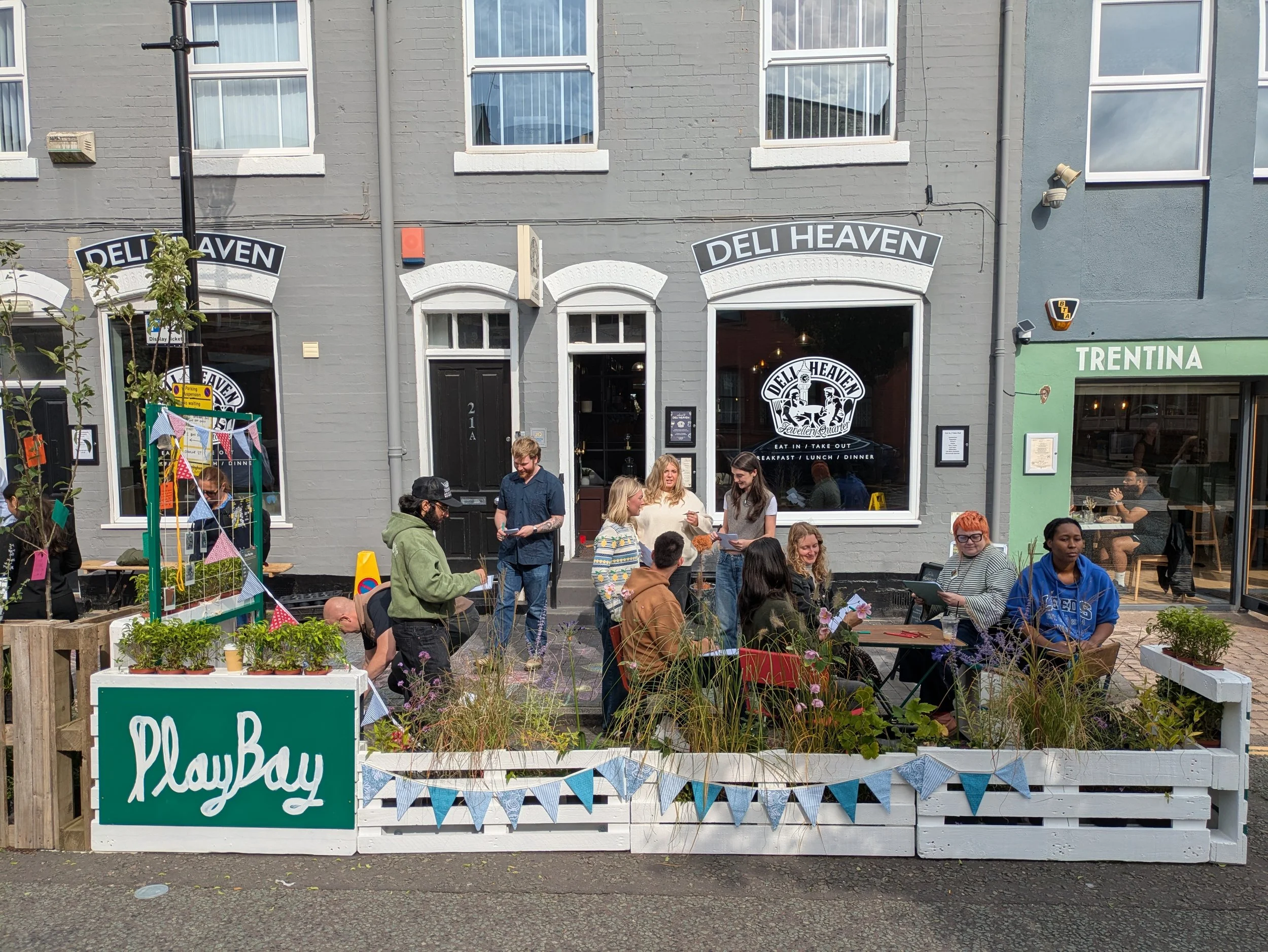 People gathering outside a shop called Deli Heaven, with a sign advertising breakfast, lunch, and dinner. A small play area with bunting and potted plants is in the foreground. There is also a green storefront named Trentina to the right.