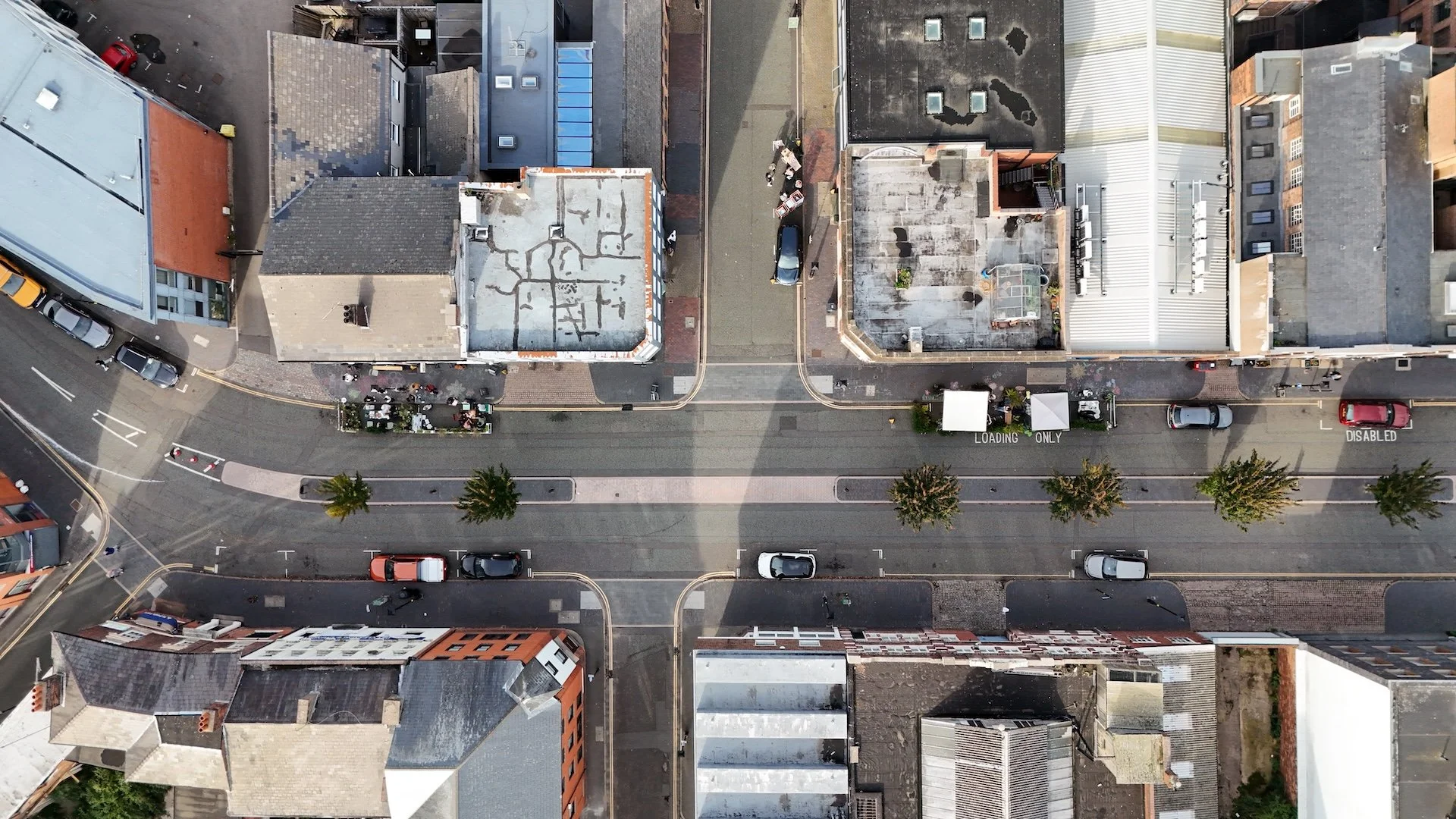 An aerial view of a city street with parked cars, buildings, and trees, showing a clear road with marked lanes and some outdoor seating areas.