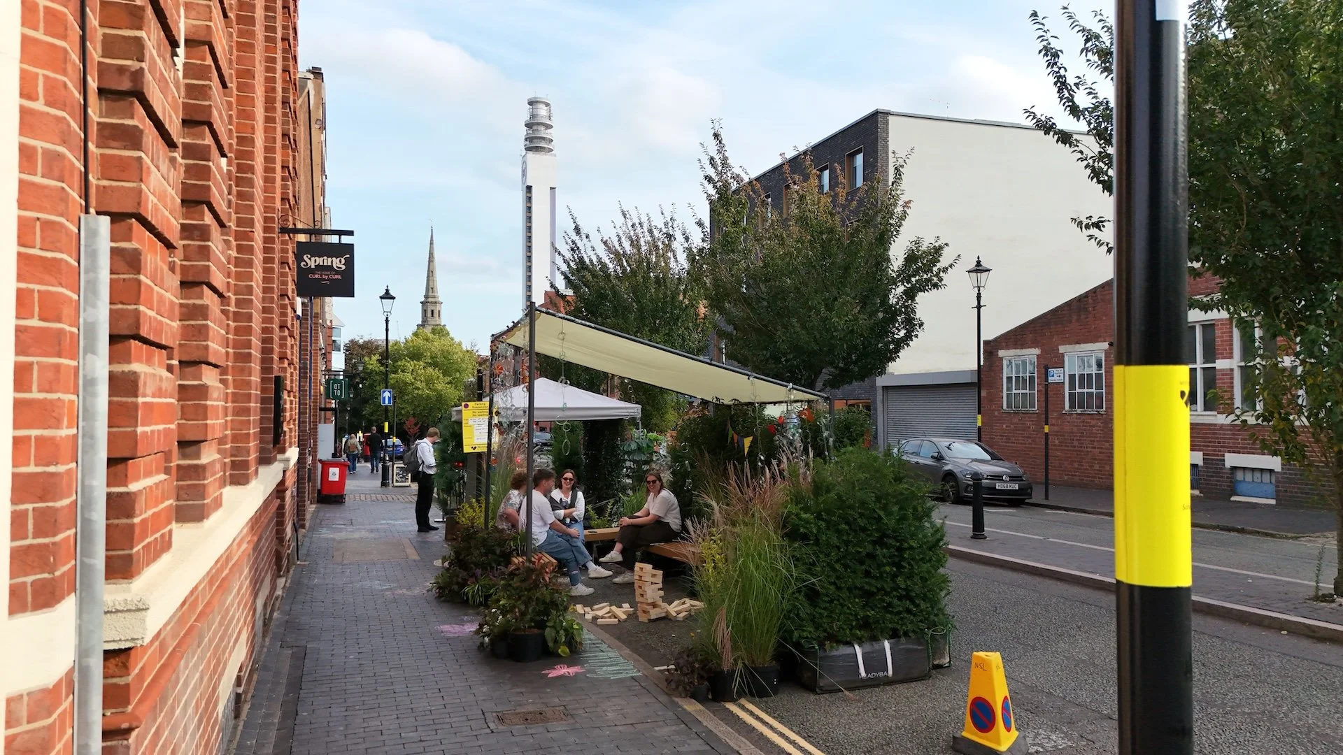 People sitting and chatting under a white canopy on a city sidewalk with brick and modern buildings, a black street lamp, and a parking lot with cars in the background.