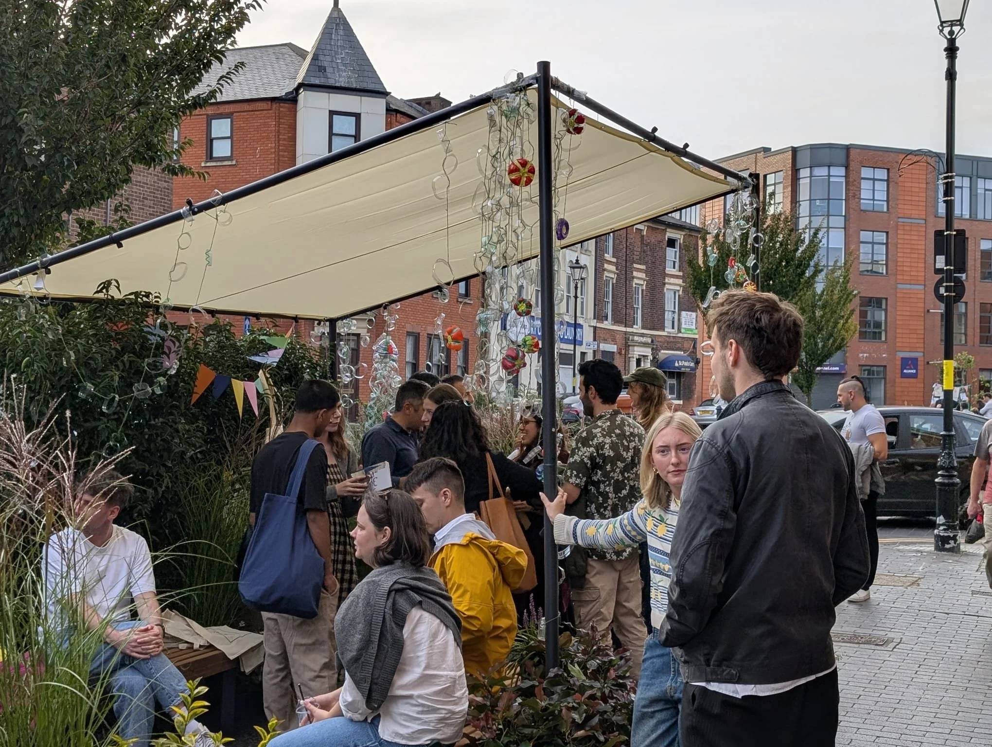 A crowd of people gathered outdoors under a canopy decorated with hanging glass ornaments and paper lanterns, with buildings and trees in the background.