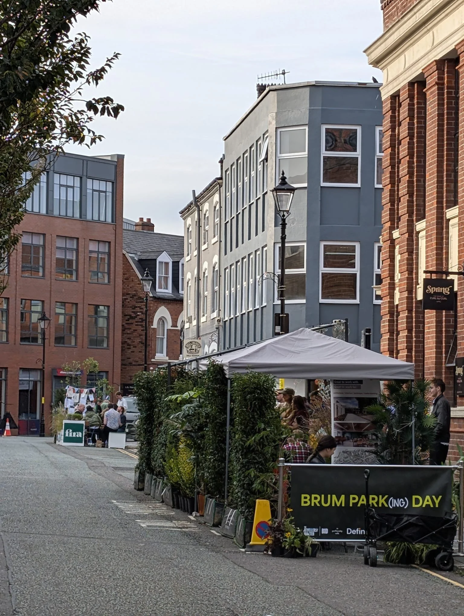 A street scene during Brum Park Day with people sitting at a table, tents, and a sign that reads 'BRUM PARK DAY' in the foreground. Buildings and streetlights are visible in the background.
