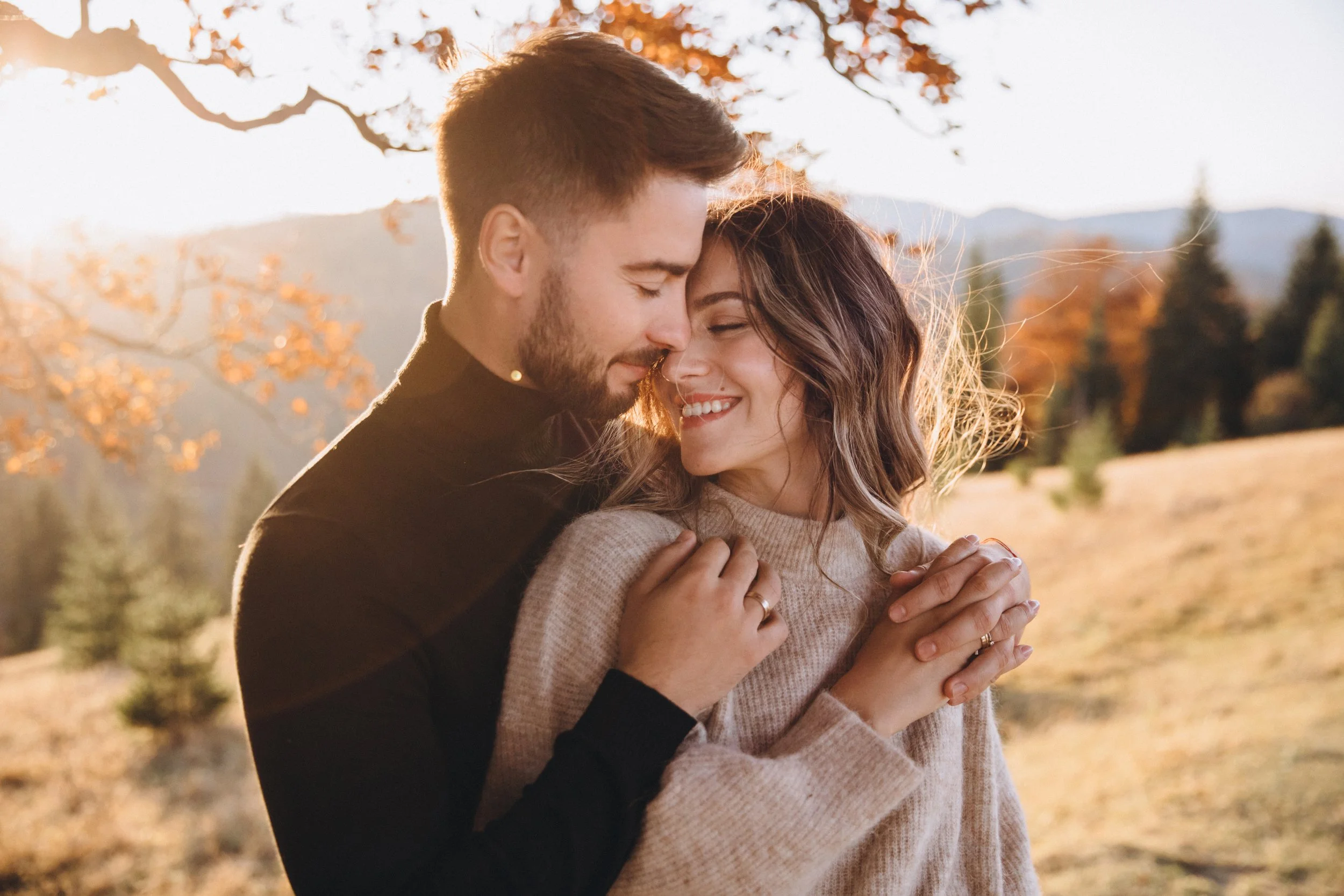 A smiling couple sharing a close embrace outdoors during sunset, surrounded by fall foliage and distant mountains.