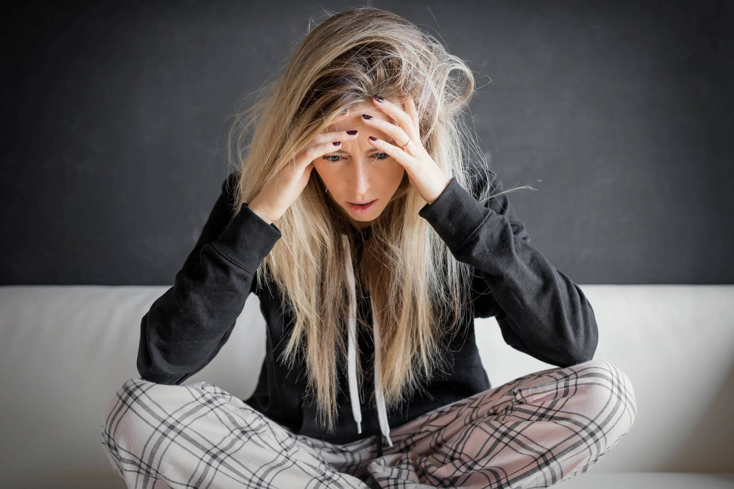 A woman sitting on a white couch with her legs crossed, holding her head in her hands with a distressed expression.