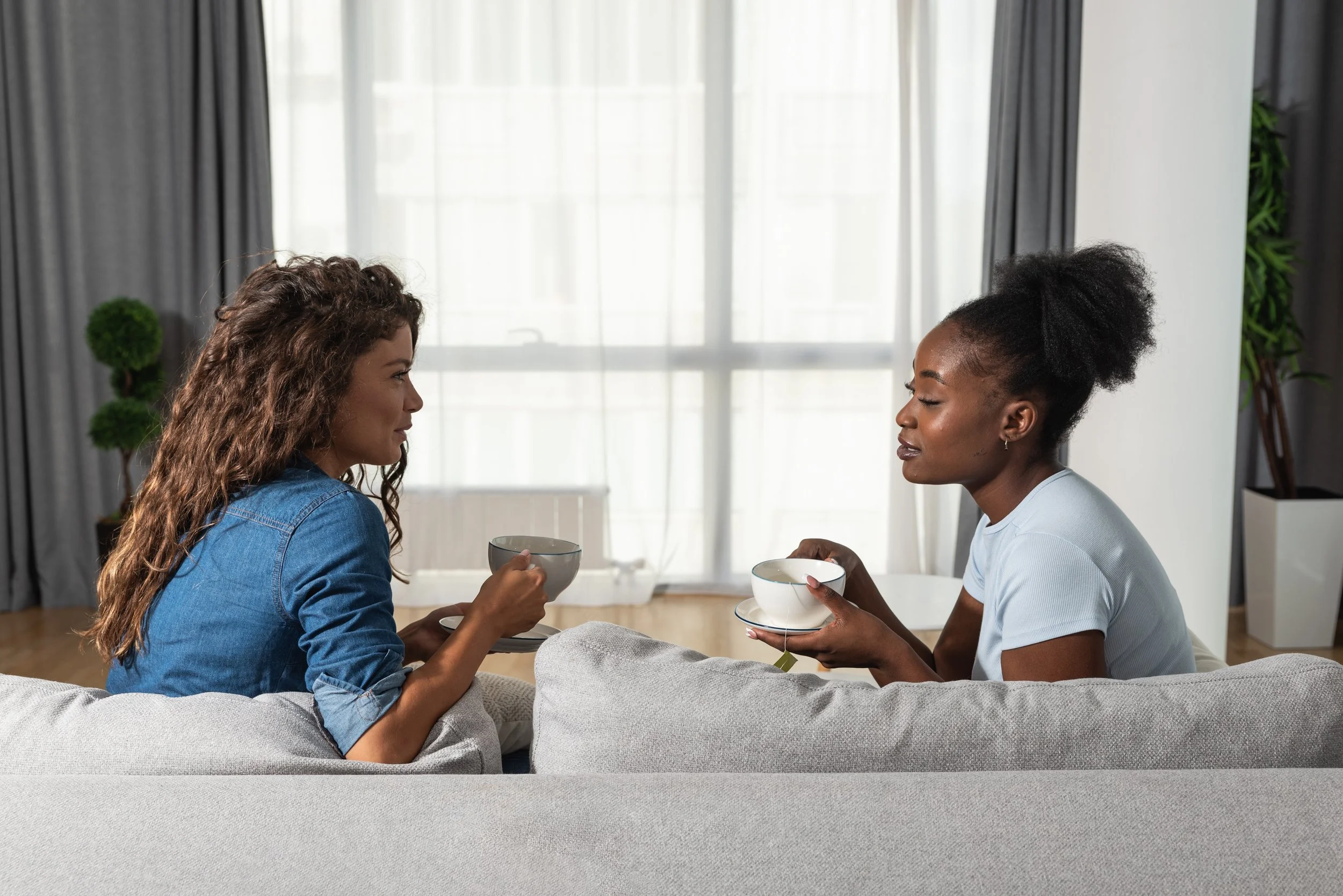 Two women sit on a couch, facing each other, each holding a teacup and saucer, engaging in conversation in a bright living room with large windows and curtains.
