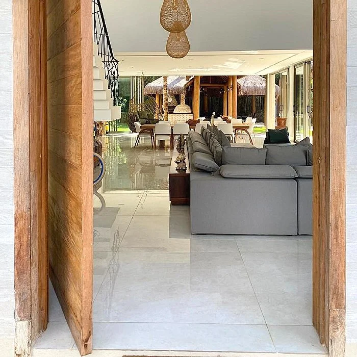 Sala de estar moderna com sofá cinza, mesa de centro de madeira, e espaço de jantar ao fundo com mesa e cadeiras, visível através de uma porta de madeira aberta.