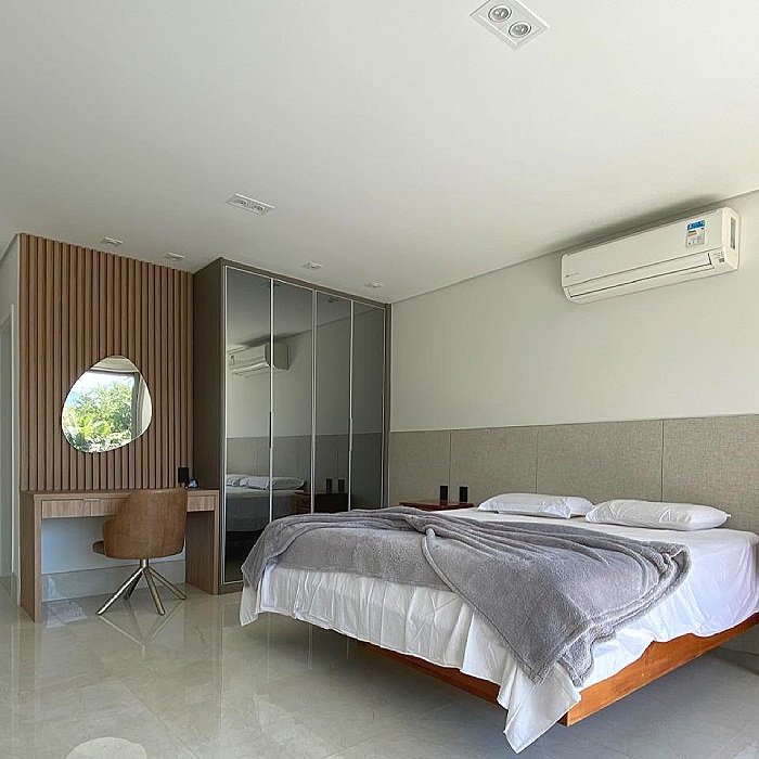 Quarto de hotel com cama de casal, cortina de vidro, espelho redondo no painel de madeira, cadeira de escritório, ar-condicionado na parede, paredes brancas e piso claro.