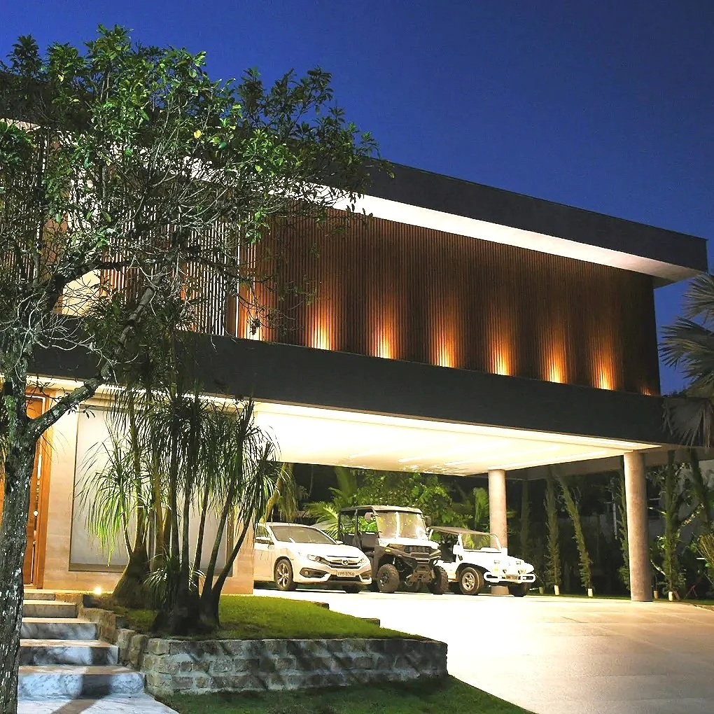 Residência moderna iluminada à noite, com fachada de madeira, carros estacionados na frente, árvores e jardim bem cuidado.