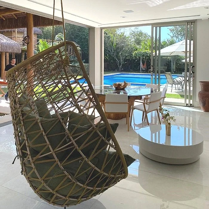 Sala de estar com móveis modernos, mesa de jantar, poltronas e uma cadeira de balanço de vime, com vista para uma piscina ao ar livre.