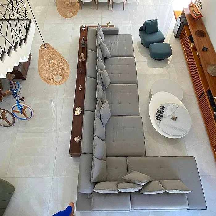 Sala de estar com sofá cinza em forma de L, mesa de centro de mármore, cadeira azul, estante de madeira, luminária de fibra natural, bicicleta ao lado da escada, e decoração de conchas na mesa