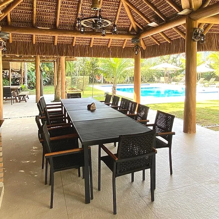 Área de lazer com mesa de jantar grande, cadeiras de madeira, cobertura de madeira quechua, piscina ao ar livre, jardins e guarda-sóis ao fundo.