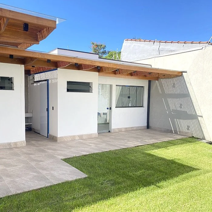 Imagem de um espaço ao ar livre com gramado e uma área de terraço pavimentada, junto a uma casa moderna com paredes brancas, janela grande, porta de vidro e teto de madeira com sombra.