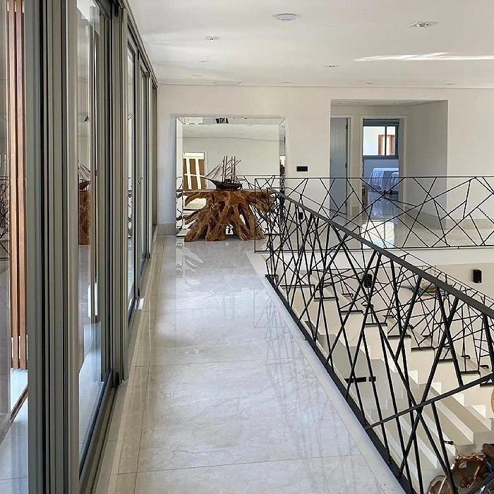 Corredor de um apartamento com piso de mármore, paredes brancas, janelas de vidro ao longo do lado esquerdo e uma escada com grade de ferro dourada. Ao fundo, uma mesa de madeira rústica com um barco em miniatura e uma cadeira ao lado.