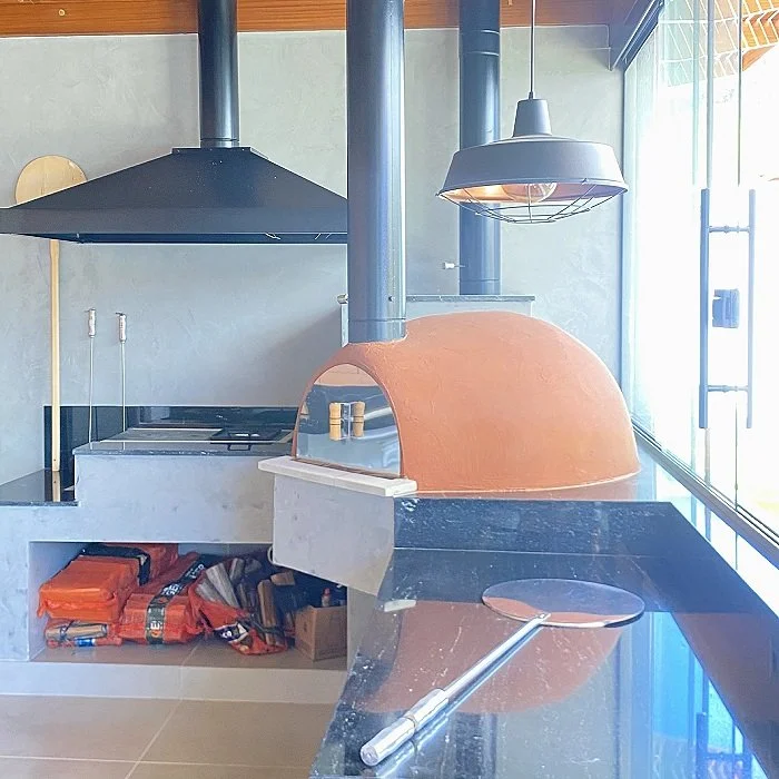 Forno de pizza de tijolo com chaminé, fogão de bancada, utensílios de cozinha, iluminação pendente e exaustor em uma cozinha bem iluminada