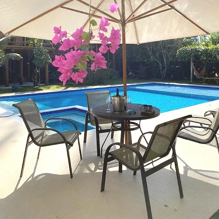 Mesa de jantar com cadeiras ao lado de uma piscina, sob um guarda-sol, com vasos de plantas e uma sombra de árvore ao fundo.