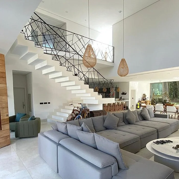 Sala de estar moderna com sofá cinza, escadas brancas, iluminação pendente de fibra natural, detalhes decorativos e área de convivência aberta com mesa de jantar.