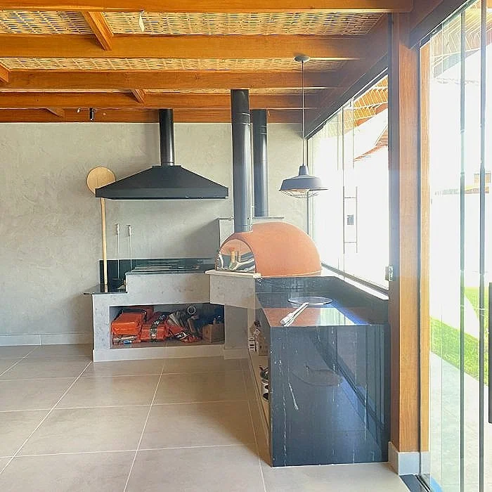 Área de churrasqueira com forno de pizza, coifa, bancada de mármore preto e cozinha integrada, no interior de uma casa com teto de madeira.
