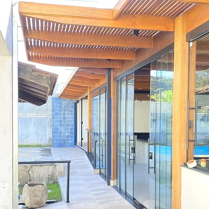 Varanda de uma casa moderna com estrutura de madeira, paredes de vidro e piso de porcelanato, durante o dia.