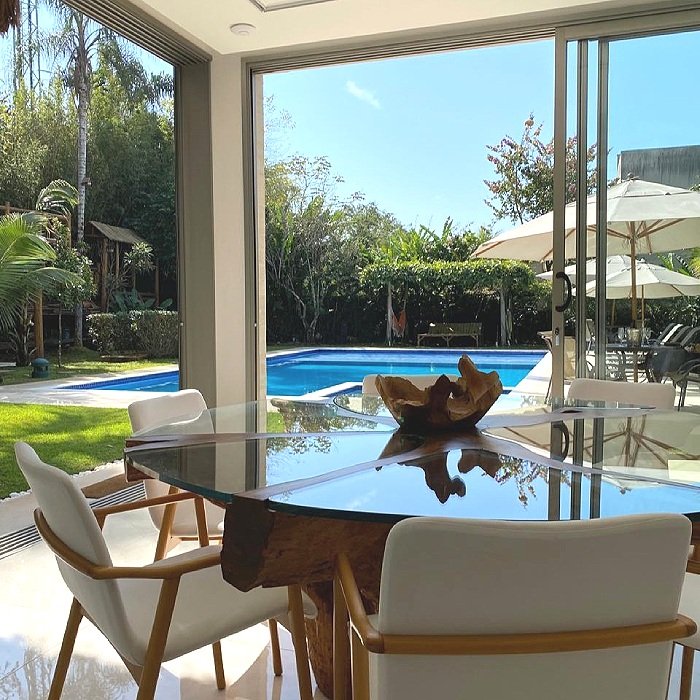 Sala de jantar com mesa de madeira com tampo de vidro, cadeiras brancas e uma vista de uma piscina ao ar livre com espreguiçadeiras, guarda-sóis e árvores ao fundo, sob céu ensolarado.