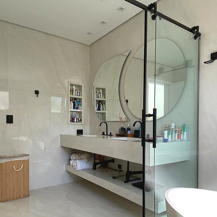 Banheiro com parede de mármore, espelhos redondidos, pia branca longa, armário com toalhas enroladas, produtos de higiene e decoração no balcão, pia preta, divisória de vidro transparente, bocal de torneira preto.