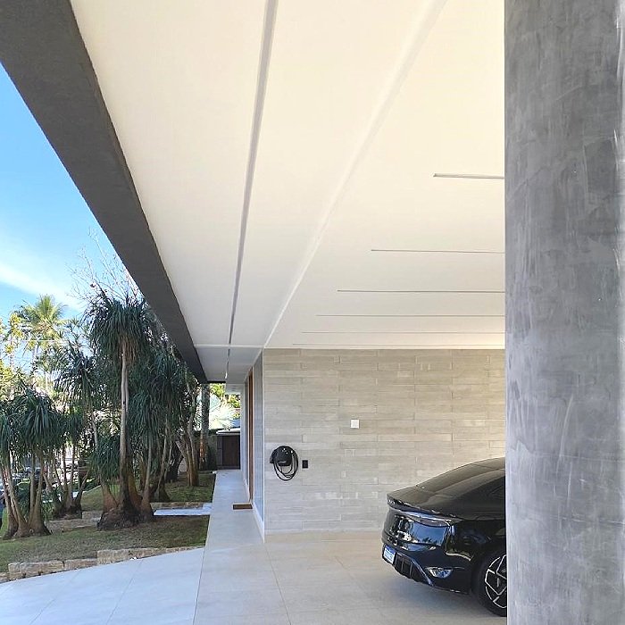 Perspectiva da varanda de uma casa moderna com teto branco, parede de pedra, carro preto estacionado na garagem, árvores ao lado e céu claro ao fundo.