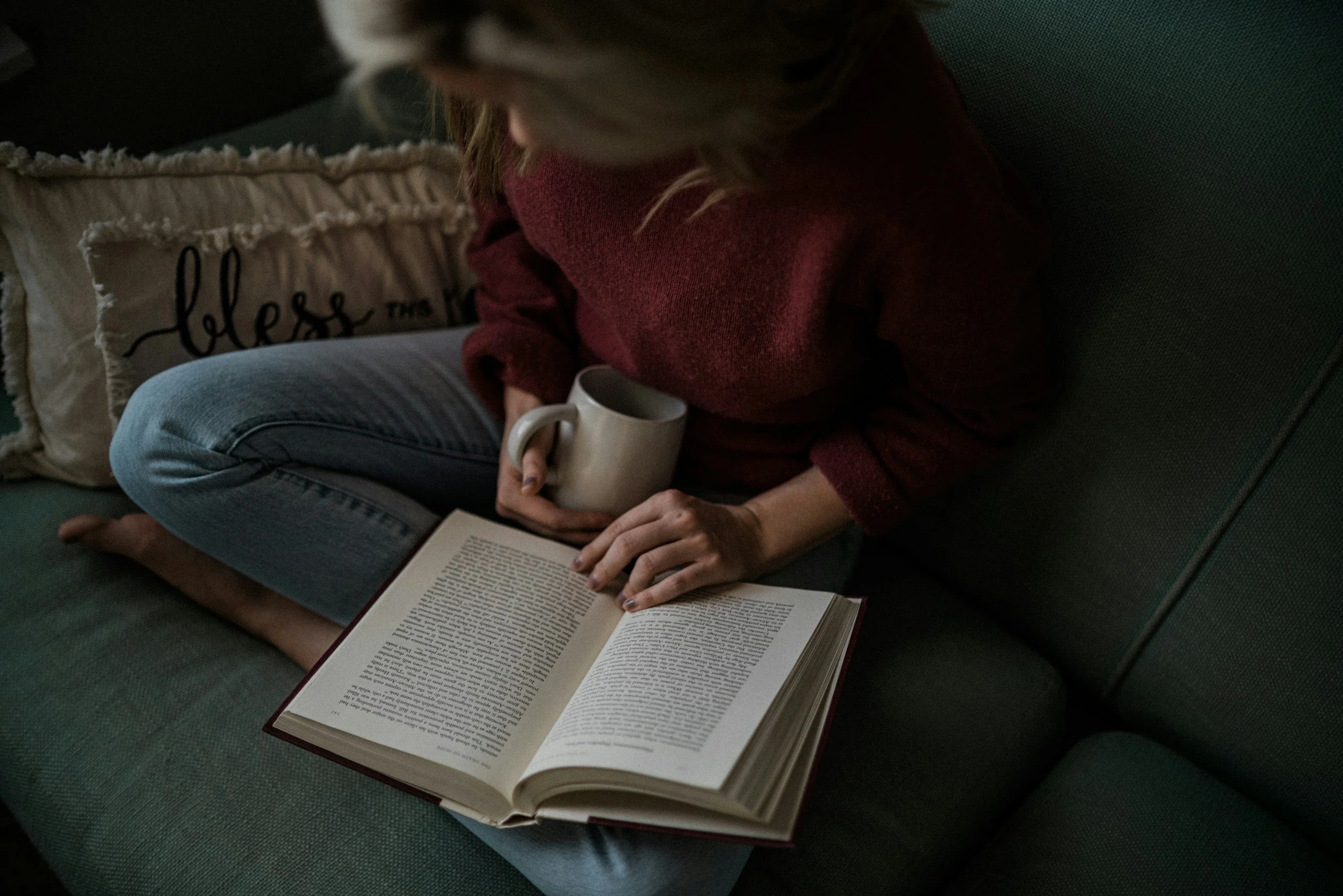 A person sitting on a green couch, reading a book, holding a coffee mug, with a decorative pillow in the background.