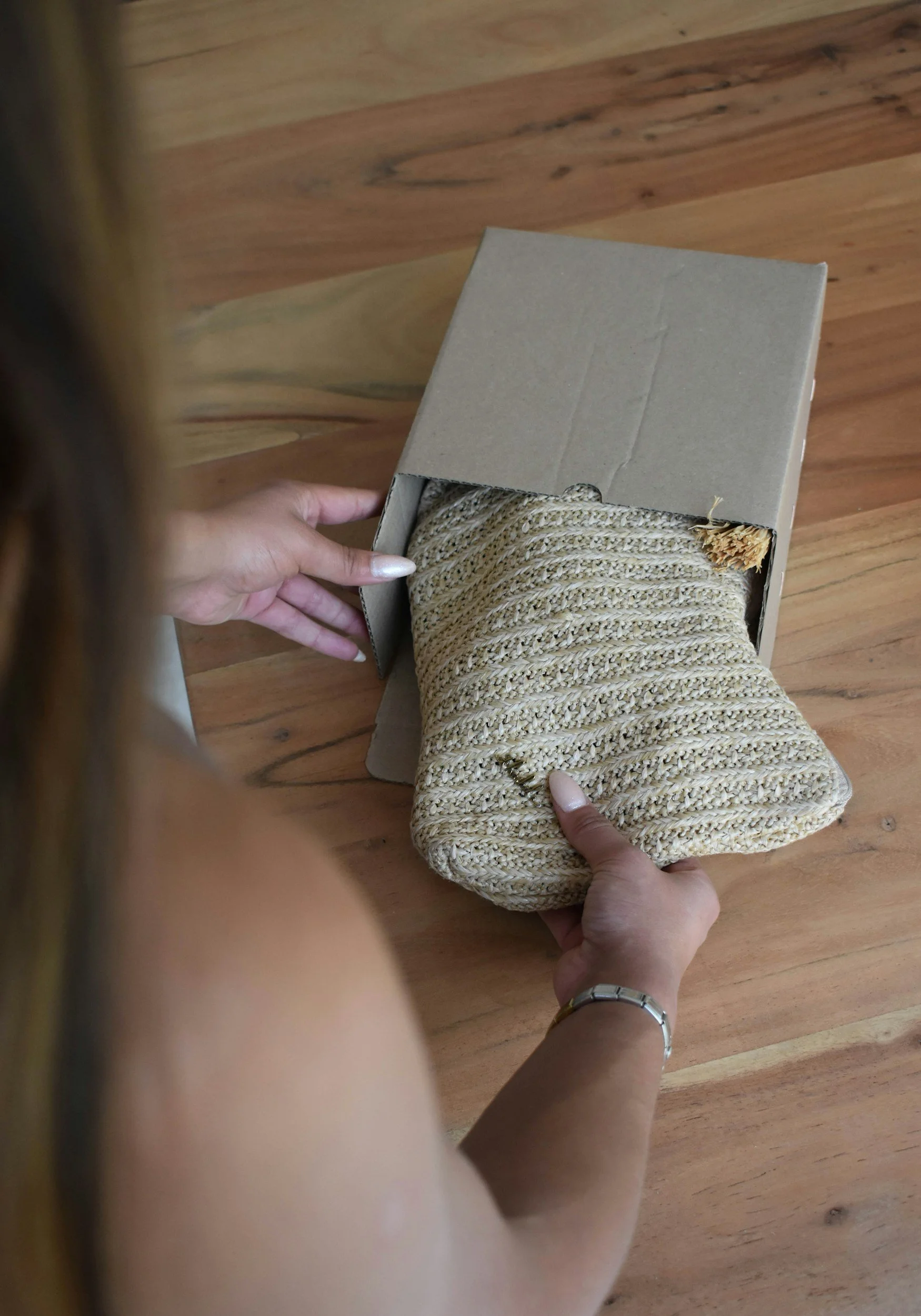 Person holding a woven beige bag and a cardboard box containing a bundle of beige rope on a wooden table.