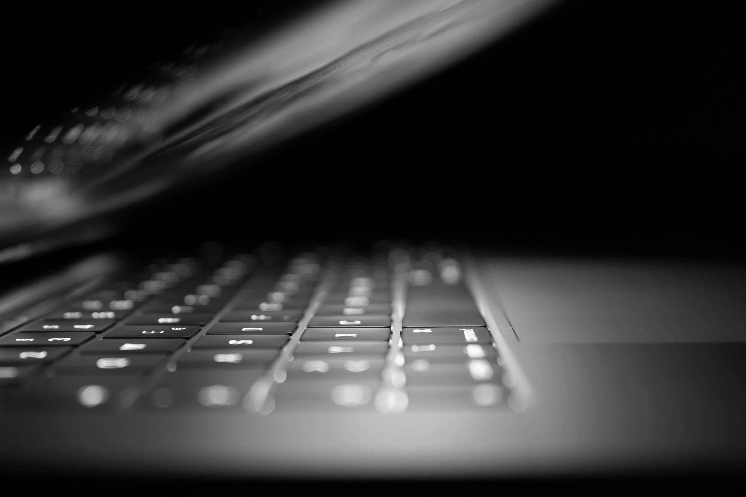 Close-up of a laptop keyboard in black and white with a focus on the keys, partially illuminated, with a blurred dark background.