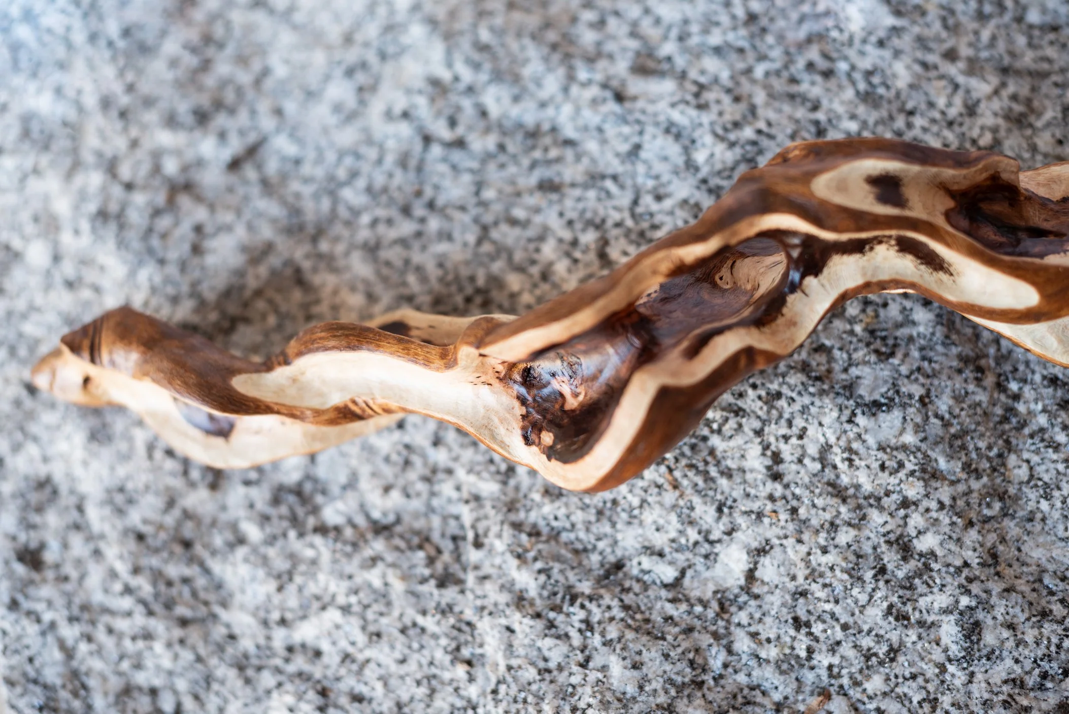 A twisted, abstract piece of wood with dark and light brown streaks resting on a grainy, speckled surface.